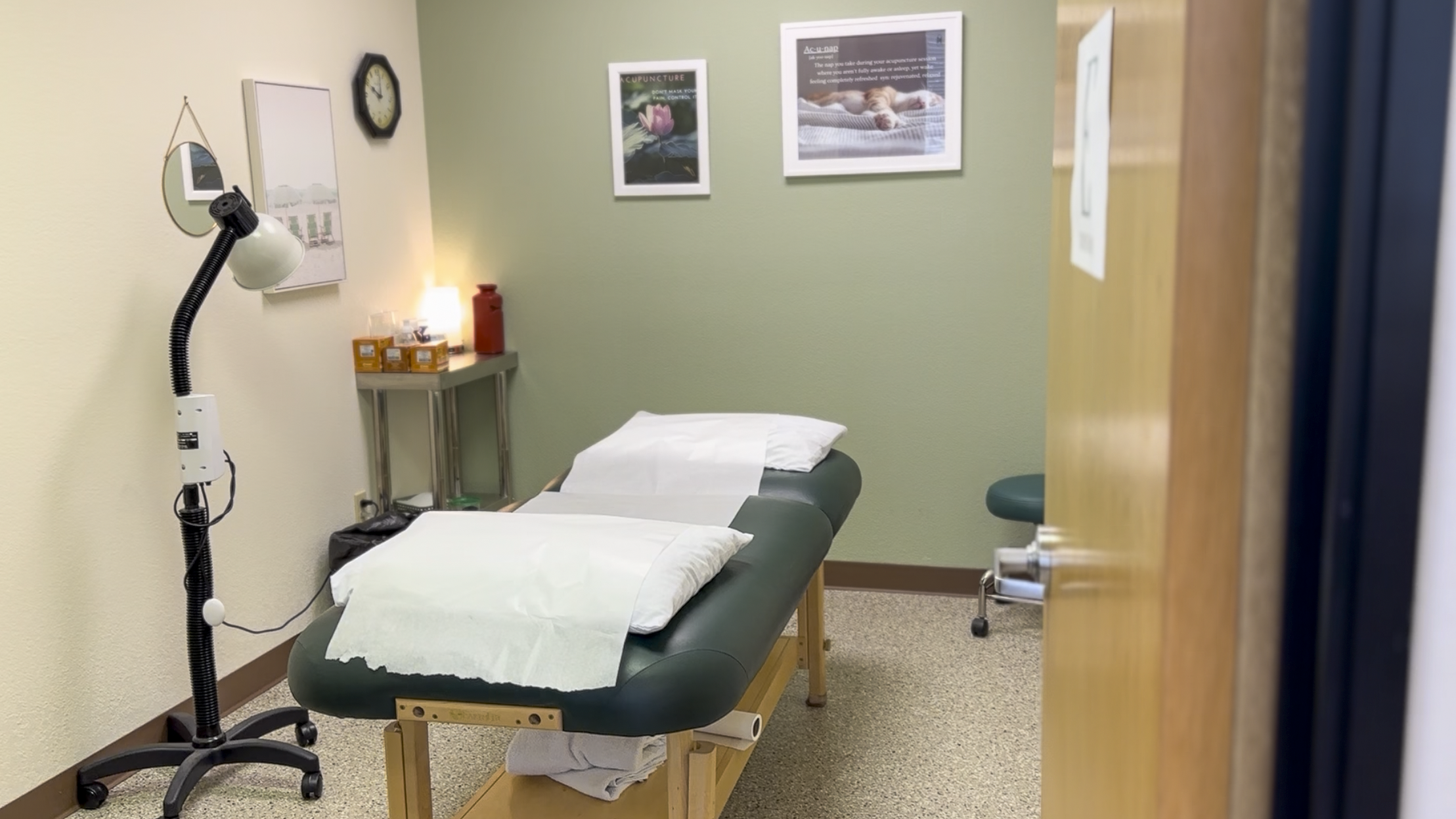 Medical examination room with treatment table, lamp, framed pictures, clock, side table, and stool.