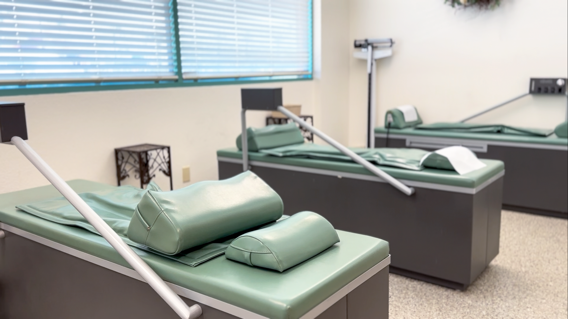 Empty hospital beds with green cushions and pillows in a medical room, with each bed equipped with a white adjustable armrest and located near windows with closed blinds.