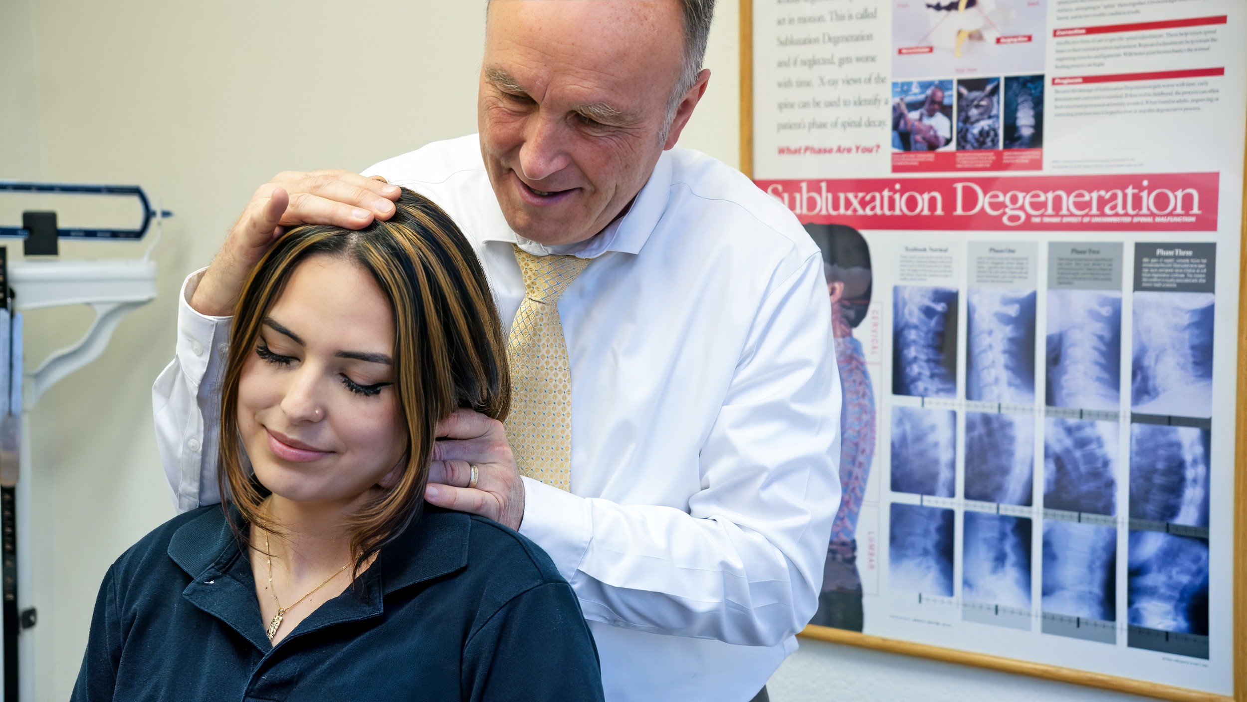 A man in a white dress shirt and yellow tie gently massages a woman's neck in a medical setting. The woman has closed eyes and appears to be relaxed. Medical posters and X-ray images are visible in the background.