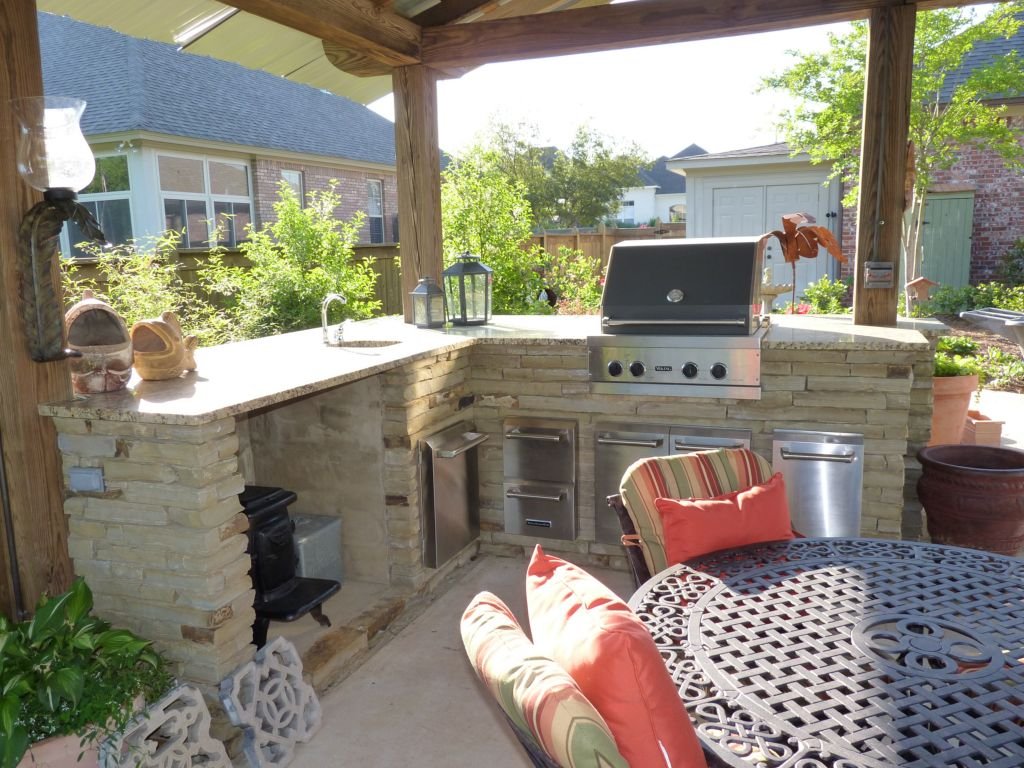 Outdoor kitchen with a built-in stainless steel grill, stone counters, and wooden pillars, surrounded by patio furniture and potted plants.