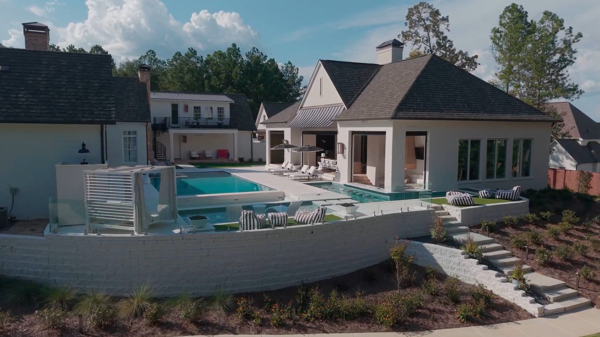 A modern house with a swimming pool in the backyard, surrounded by patio furniture and landscaping, with trees in the background.