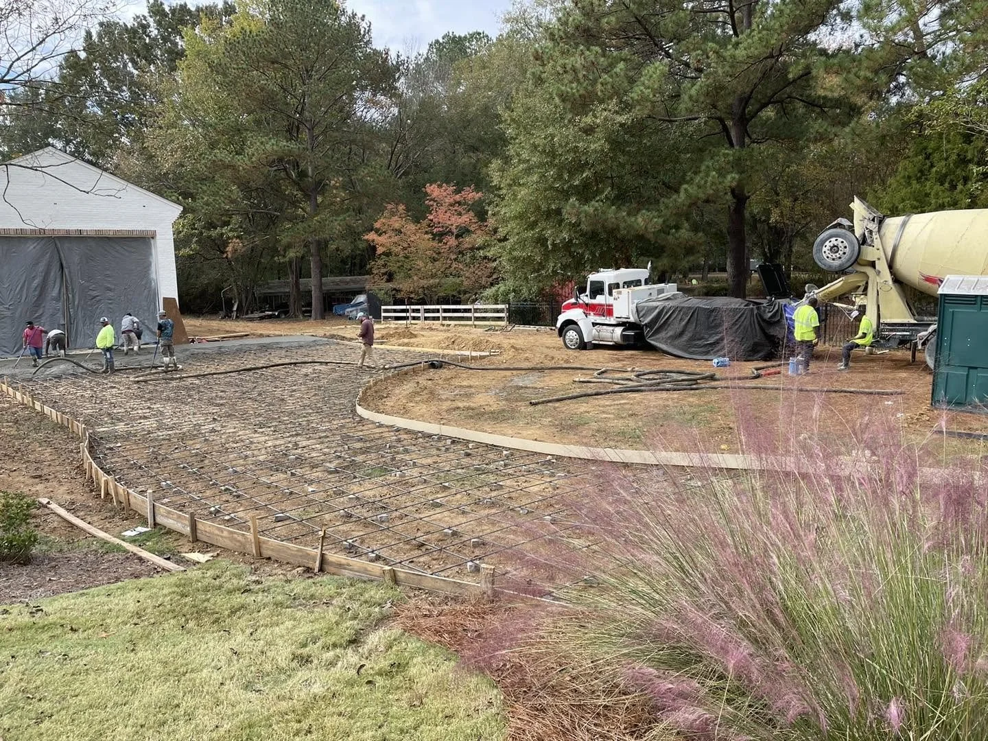 Construction workers are pouring concrete for a new sidewalk or driveway, with a concrete mixer truck and a cement pump in operation. The scene is set outdoors with trees in the background and a small grassy area in the foreground.
