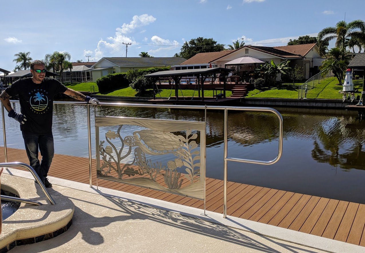 Metal art by a canal with houses in the background, a railing with a turtle design, and a wooden dock.