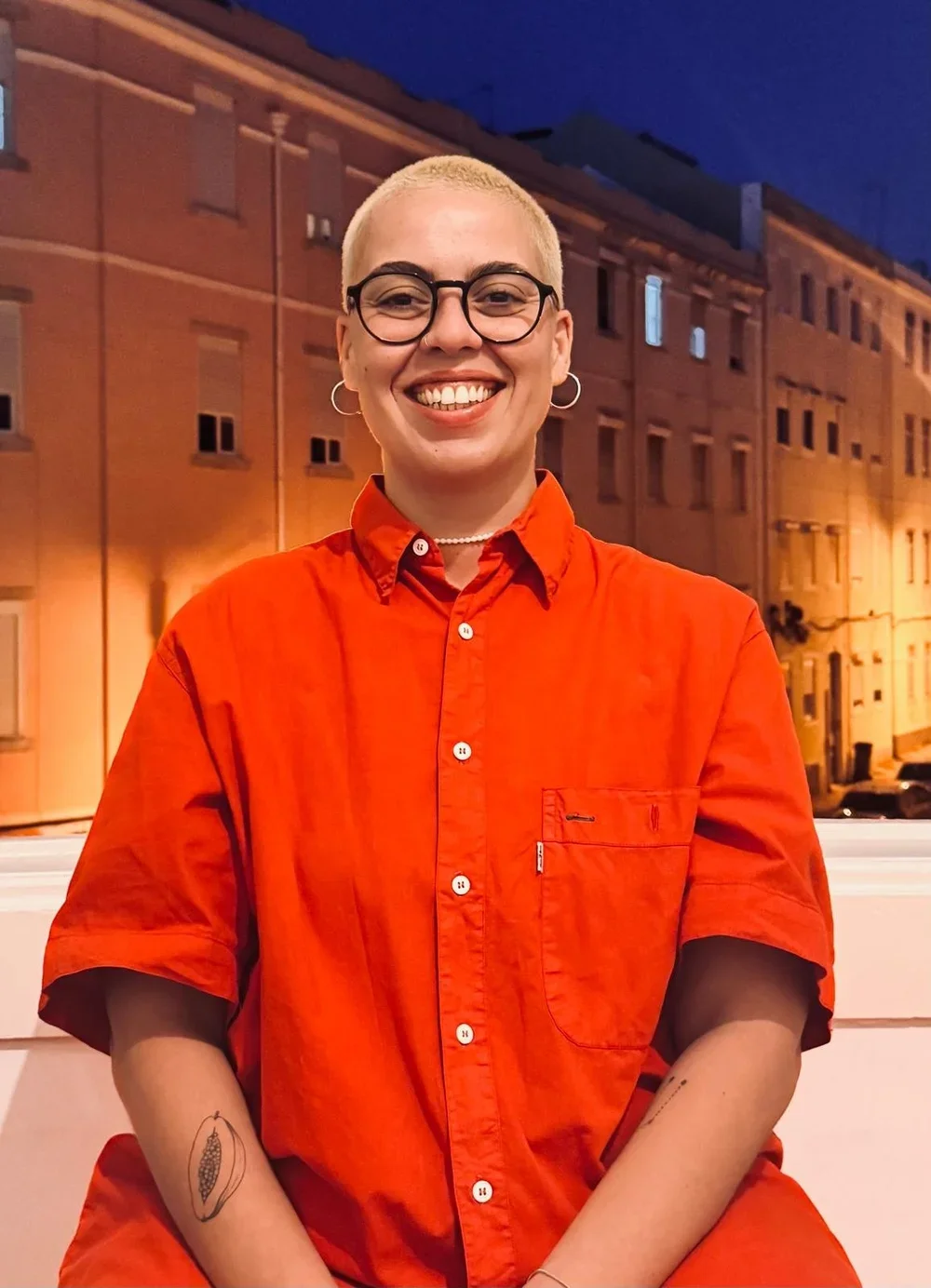 Person with short blonde hair, glasses, and hoop earrings smiling, wearing an orange button-up shirt, standing outdoors at dusk in front of a brick building with warm lighting.