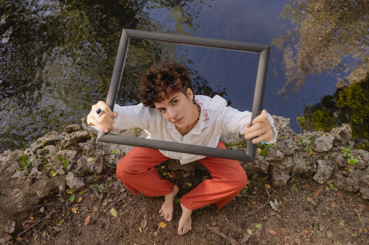 Young person with short curly hair sitting on rocks by water, holding an empty picture frame above their head, looking up.