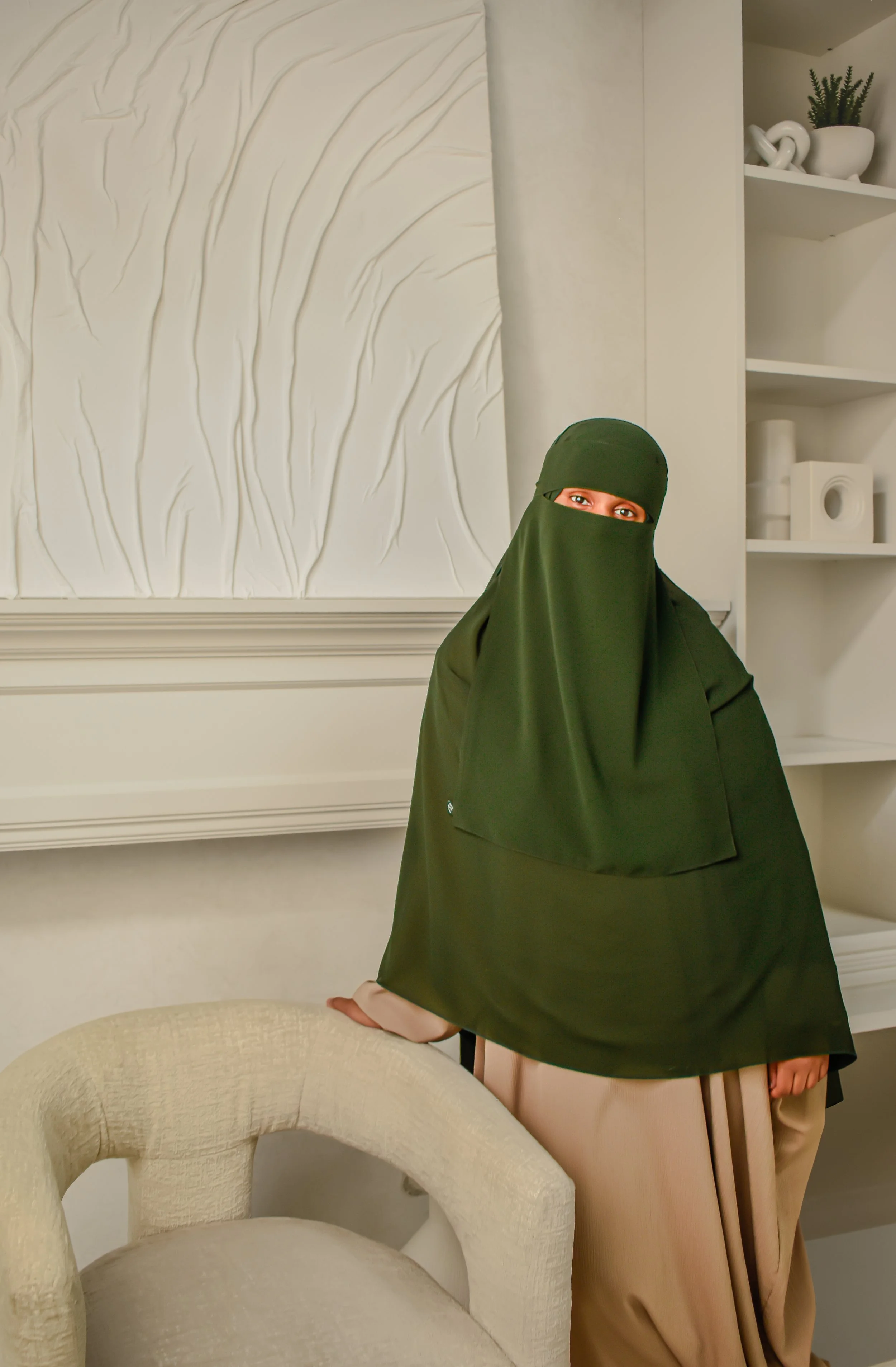 A woman wearing a dark green niqab covering her head and face, with only her eyes visible, is seated in a living room. She is wearing beige pants, and her hand rests on the beige upholstered armchair in front of her. Behind her, there is a white wall with textured artwork, and an open bookshelf with decorative items and a potted plant.