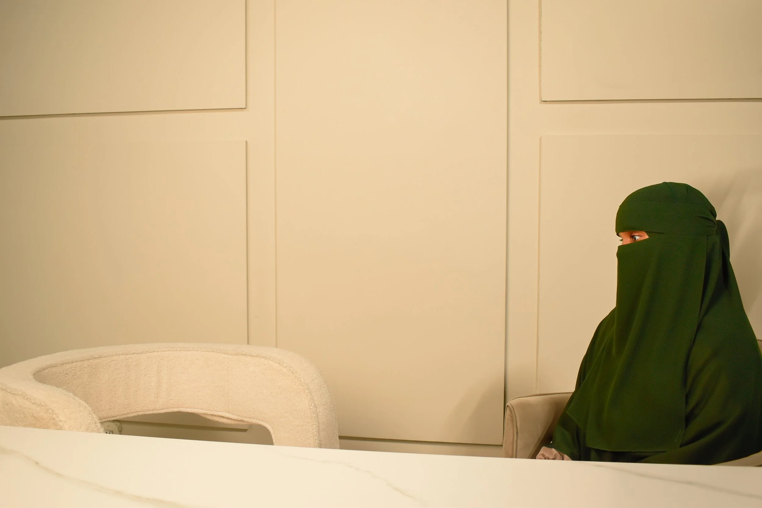 A person wearing a green niqab sitting on a beige chair in a room with cream-colored walls and white paneling. The person's face is partially visible through the niqab, revealing their eyes.