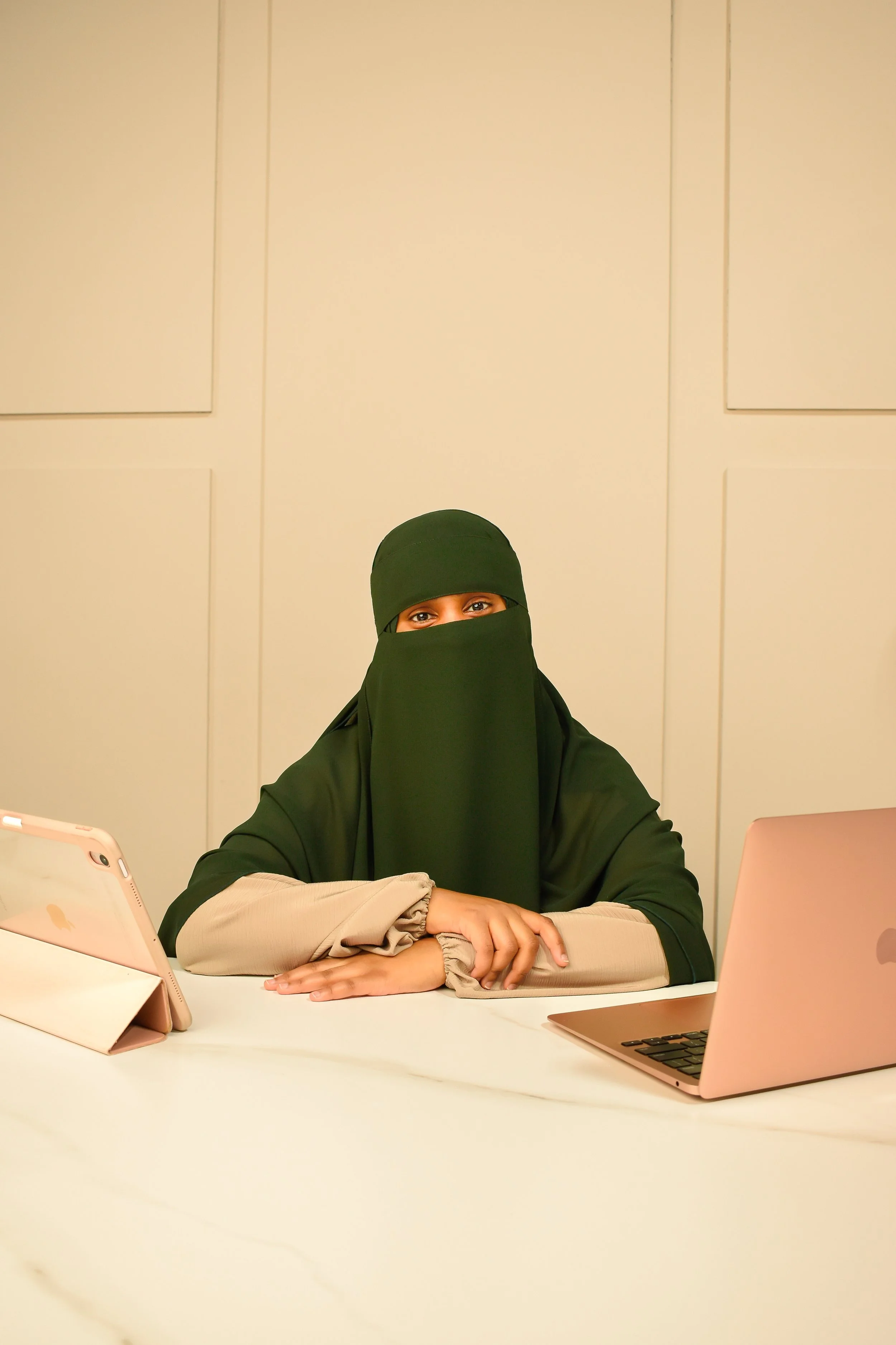 Woman wearing a black niqab and beige long-sleeve shirt sitting at a white table with a pink tablet and a rose gold laptop in an office setting.