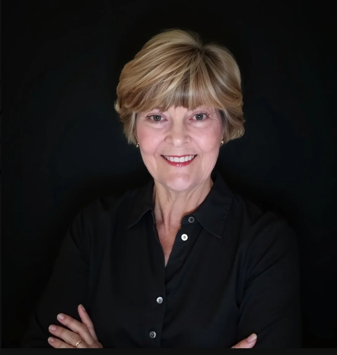 A smiling middle-aged woman with short blonde hair wearing a black button-up shirt against a black background.