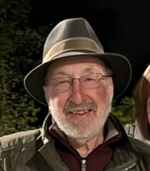 Smiling elderly man with glasses, wearing a fedora hat and outdoor jacket, standing outside at night.