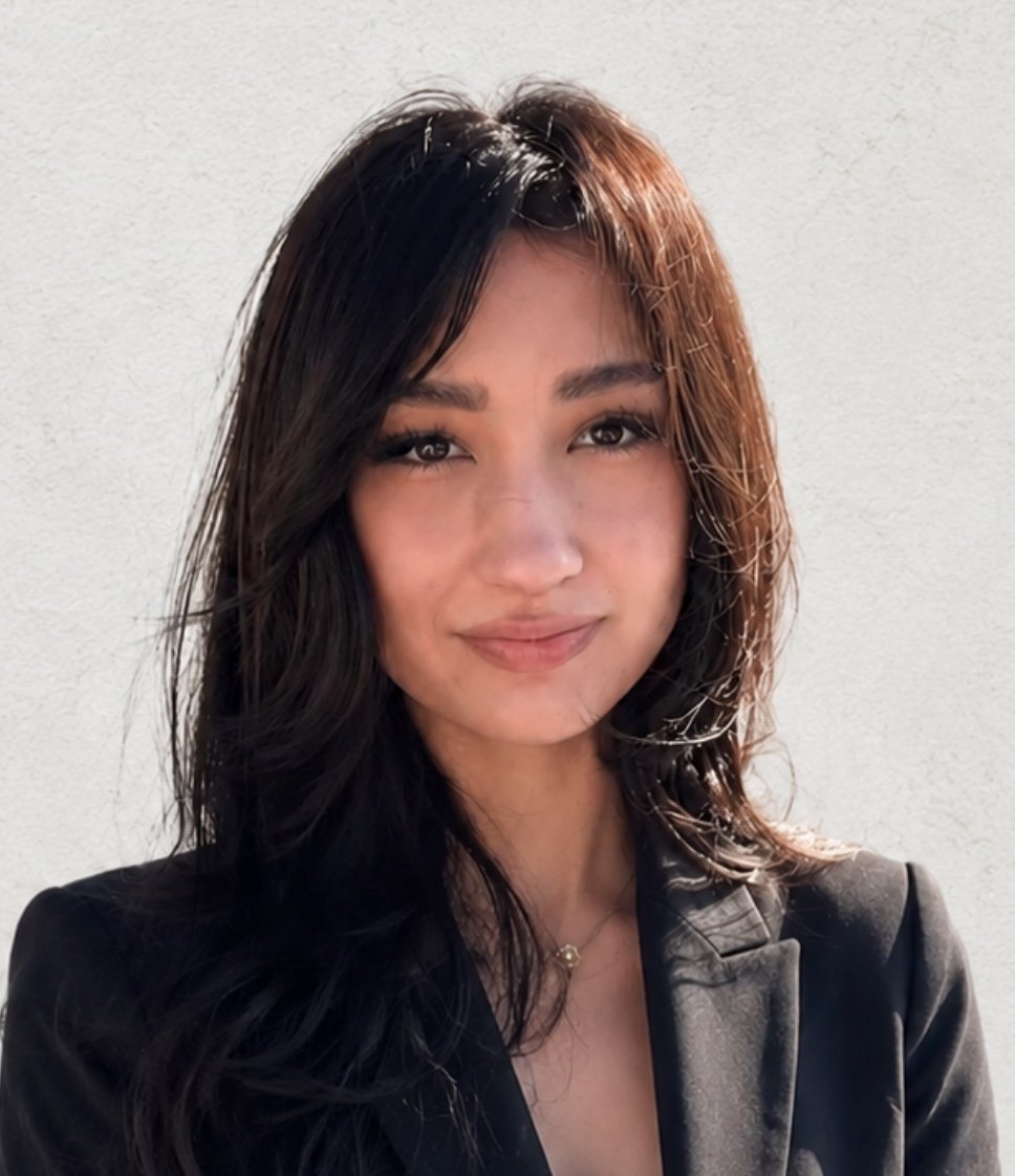 A young woman with long, wavy black hair and light brown highlights, wearing a black blazer, smiling softly in front of a white wall.