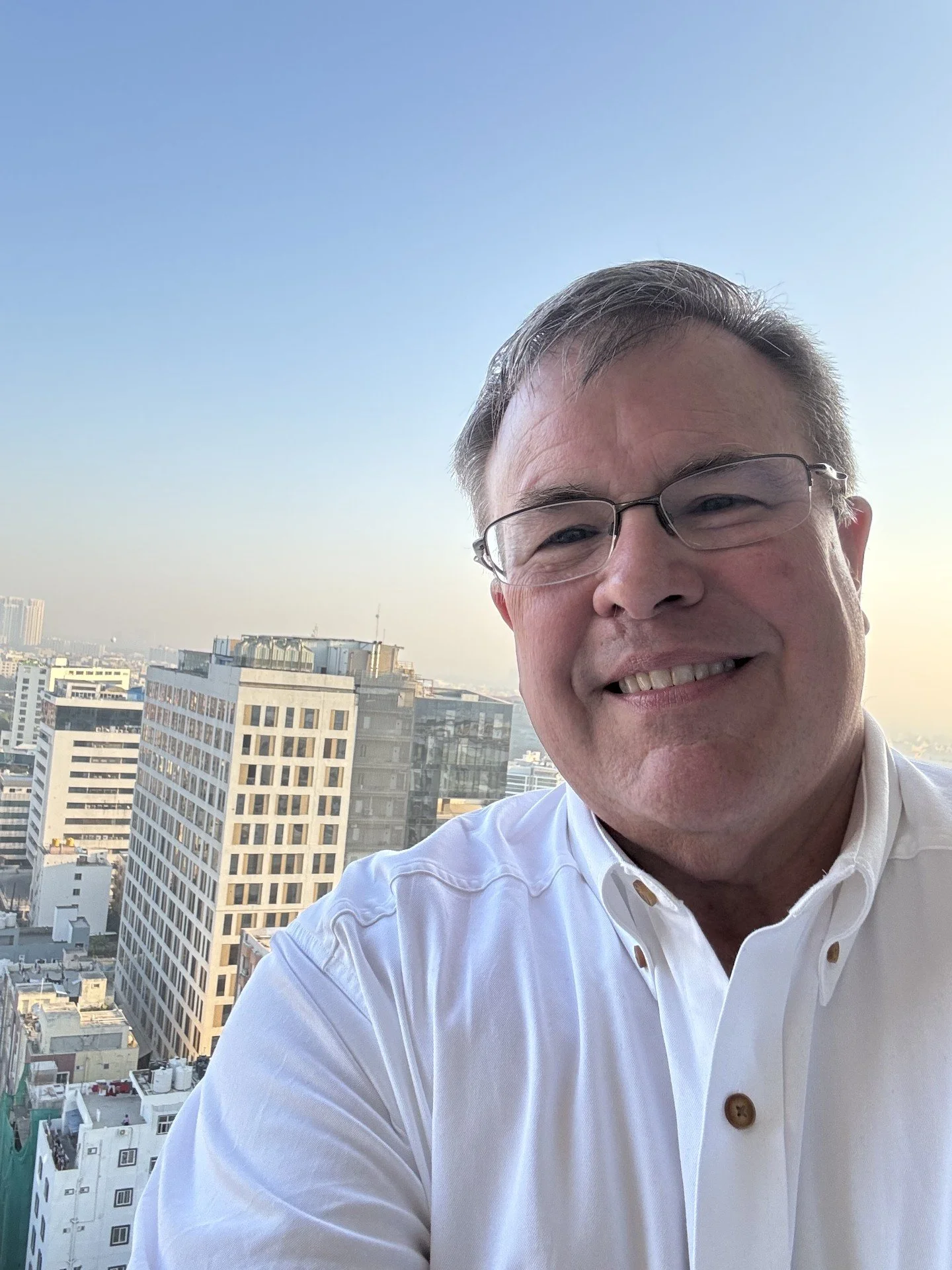 A smiling man wearing glasses and a white shirt taking a selfie on a balcony with a cityscape background of tall buildings under a clear blue sky.