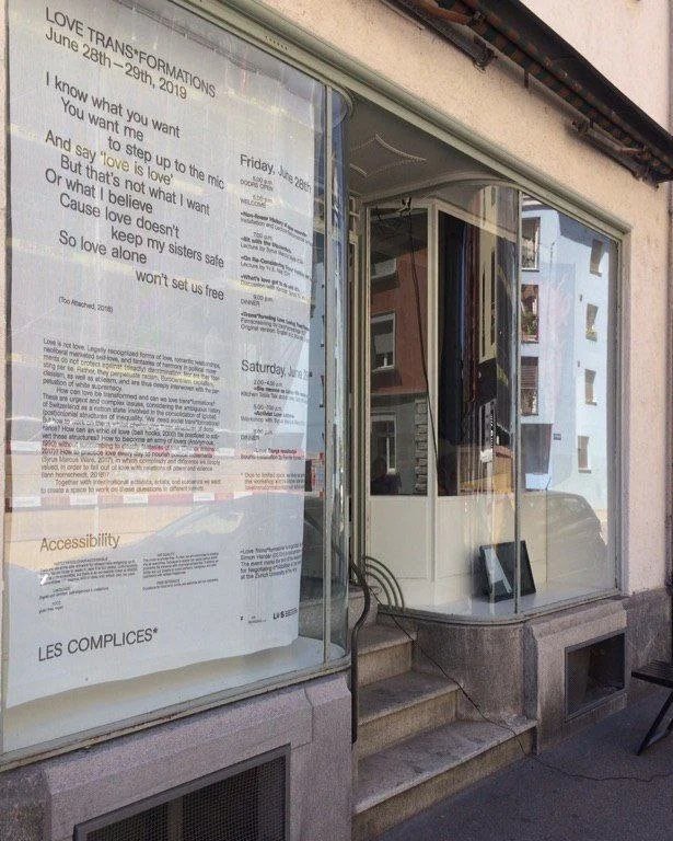 Display window with a large poster advertising Trans*formations event from June 28-29, 2019, and a small blackboard on the sidewalk outside a building with steps leading up to the entrance.