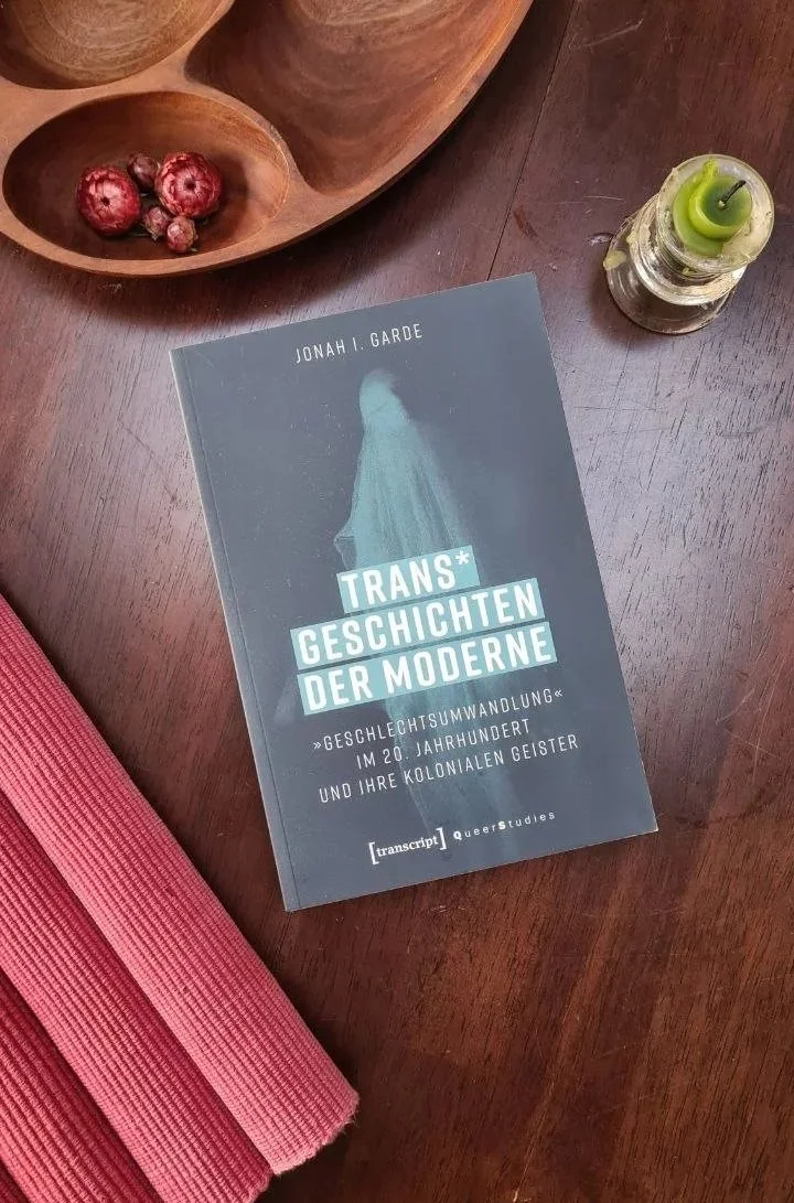 A book titled 'Trans* Geschichten der Moderne' by Jonah I. Garde is on a wooden table, along with a wooden dish with dried red berries, a small glass candle holder with a green candle, and a pink yoga mat.