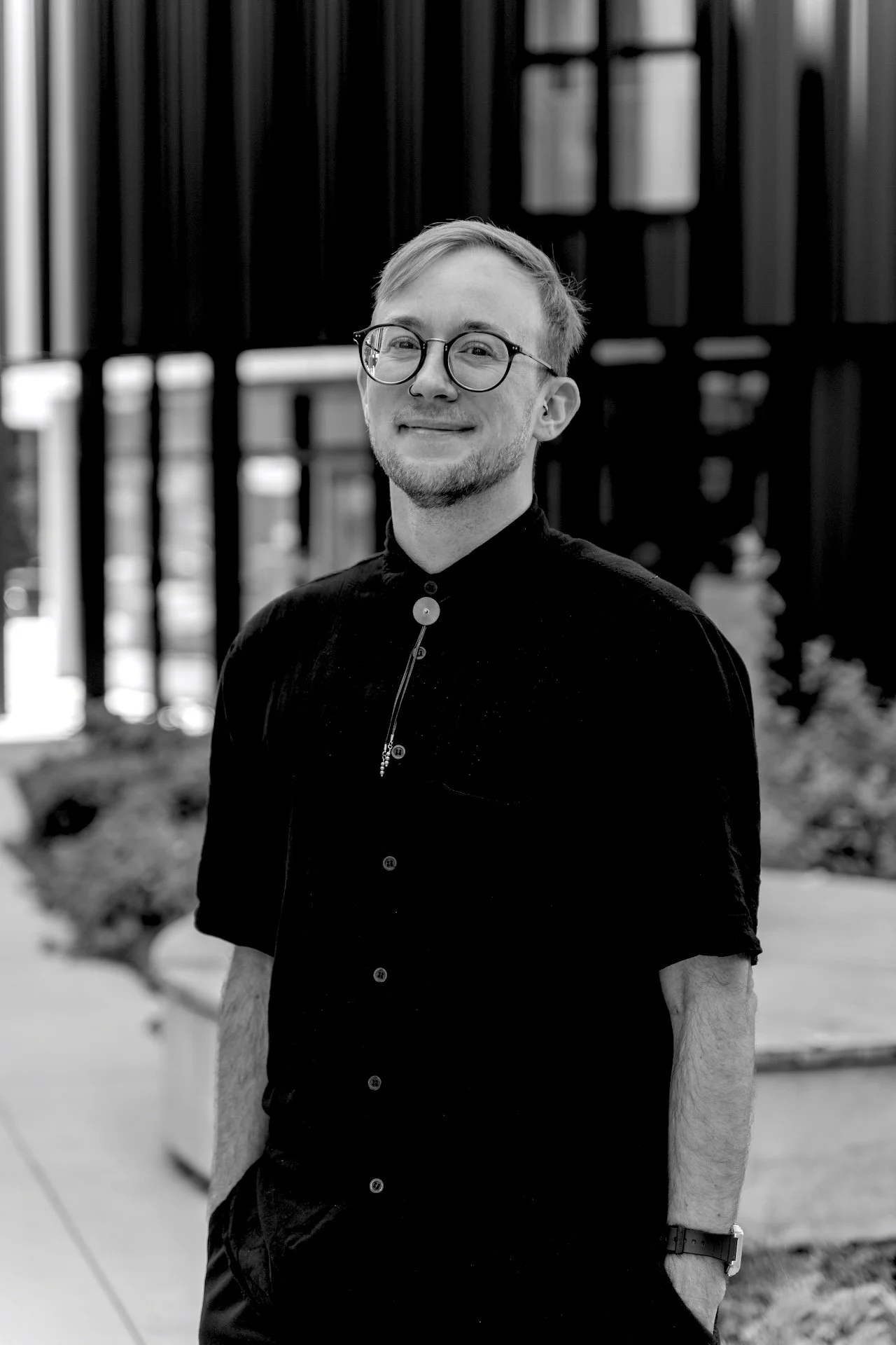 A young man with glasses and a beard, wearing a black shirt, standing outdoors with a blurred modern building in the background.