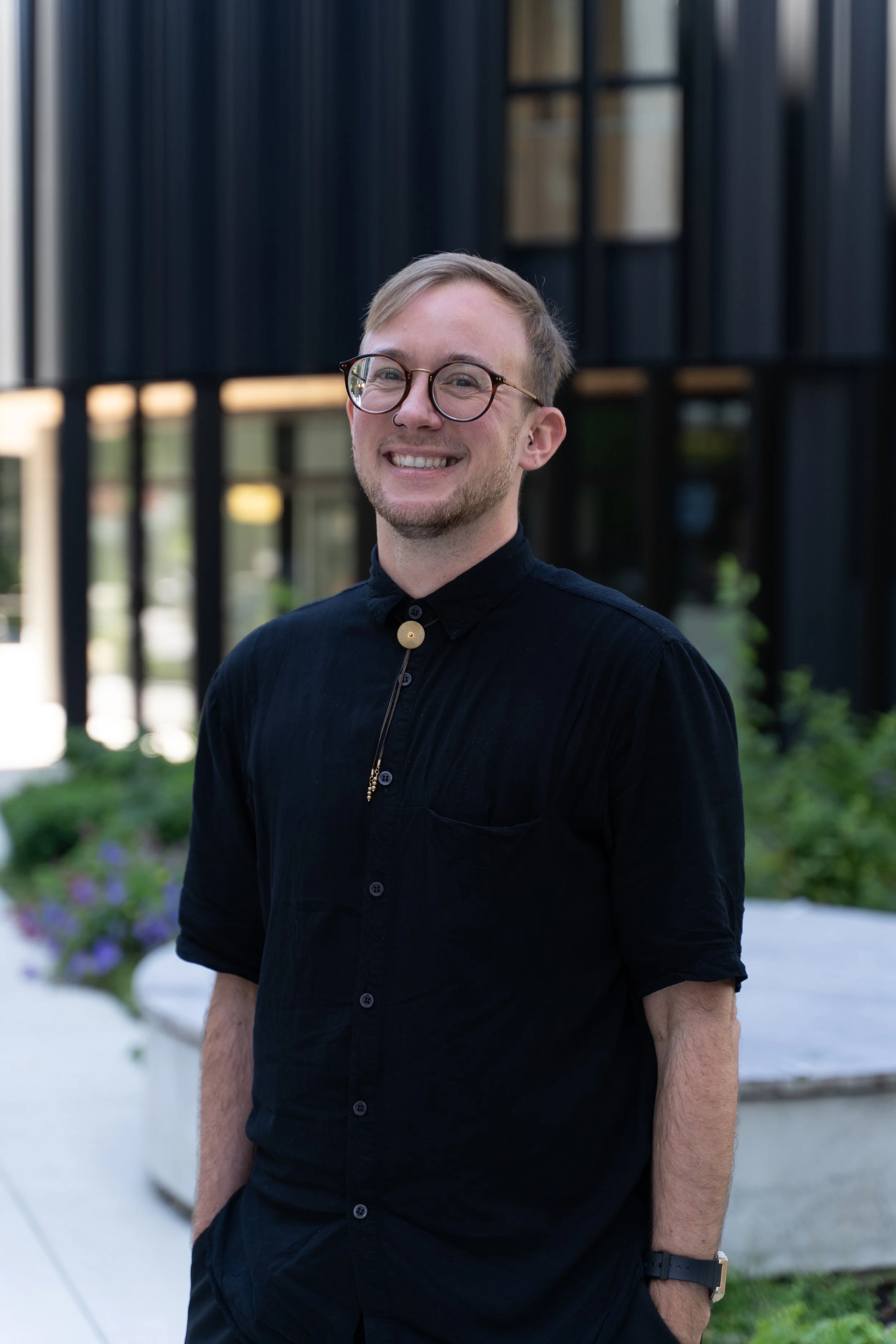 A young man with glasses and a nose ring smiling outdoors, wearing a black shirt with a pin at the collar, standing in front of a modern building with black window frames and a garden with purple flowers.