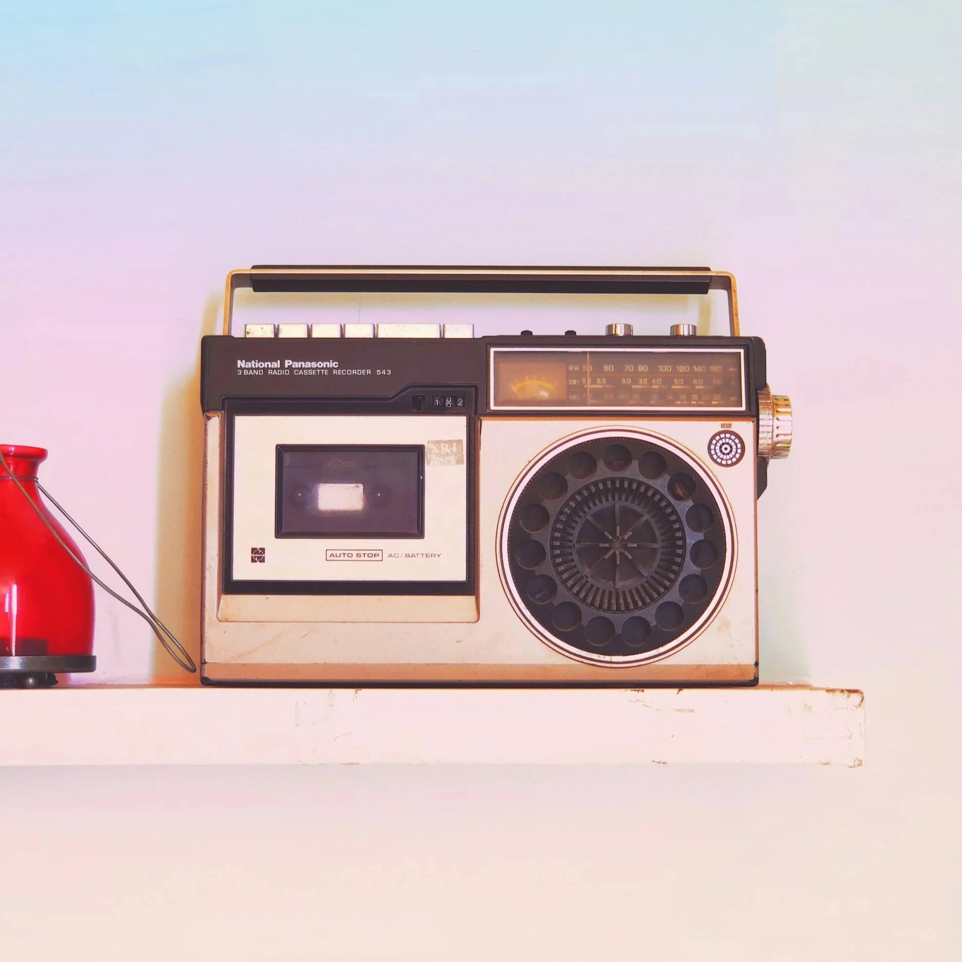A vintage portable radio cassette recorder on a white shelf with a white wall background. The radio is Black and beige with a cassette compartment, radio tuning dial, and control buttons.