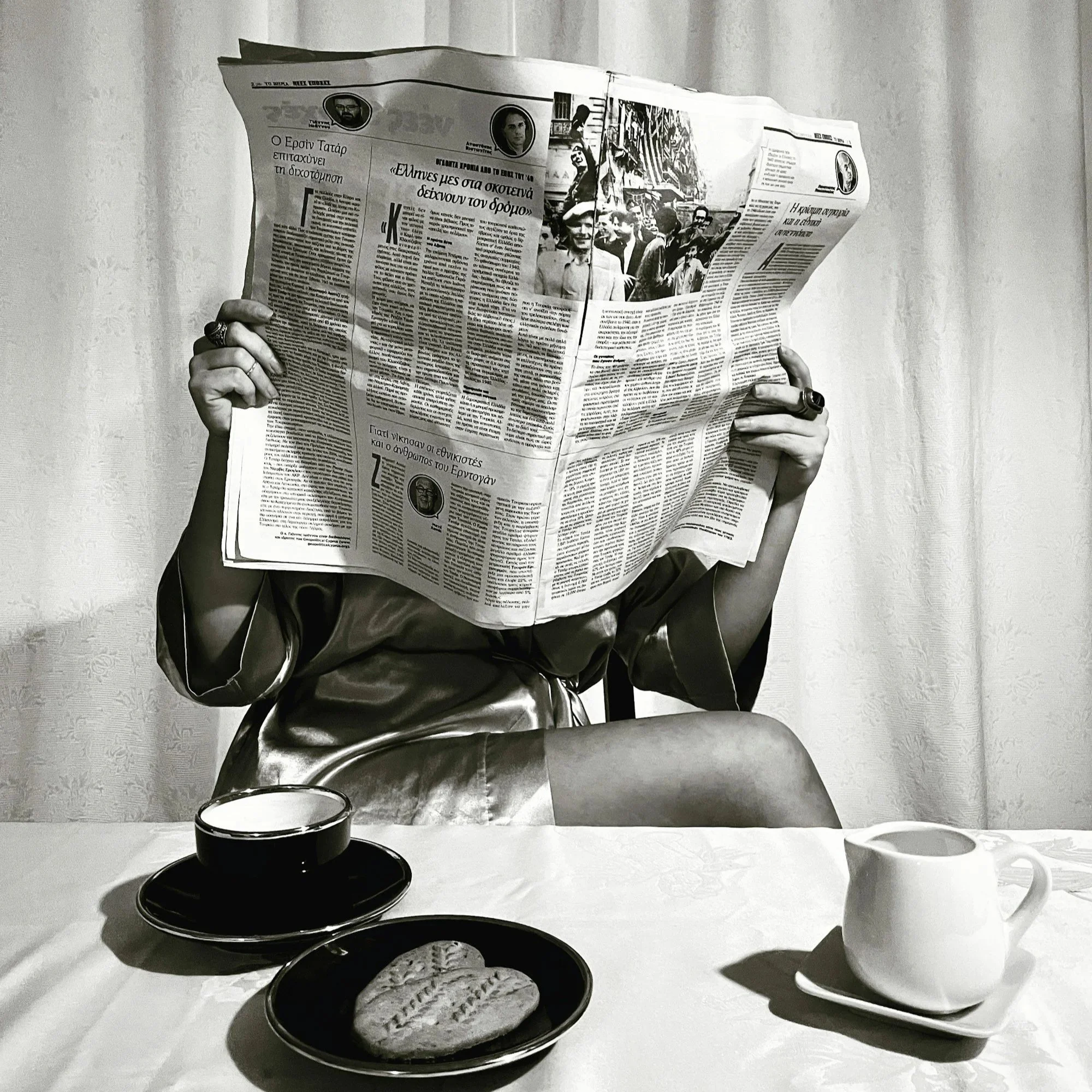 A person reading a newspaper with various images and articles, with a table in front holding a glass of dark beverage and a white bowl, in a monochrome setting.