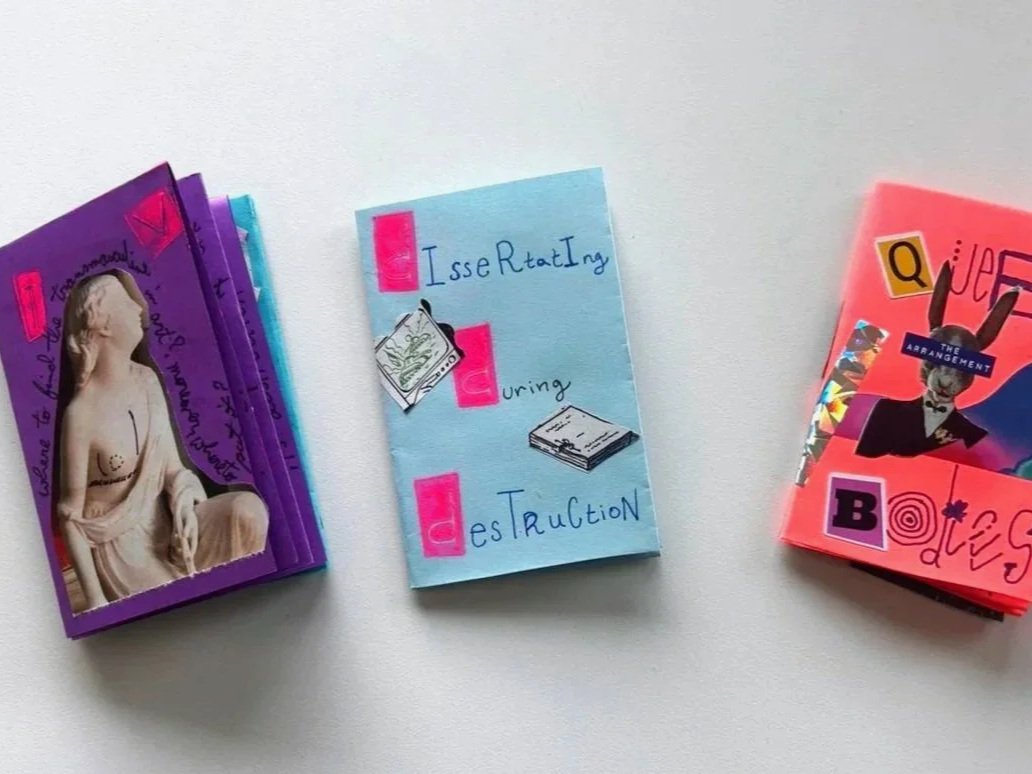 Stacked booklets with colorful covers on a white surface, including a purple booklet with a classical sculpture on the cover, a blue booklet with handwritten text and illustrations about threatening and destruction, and a pink booklet with lively graphics and text.