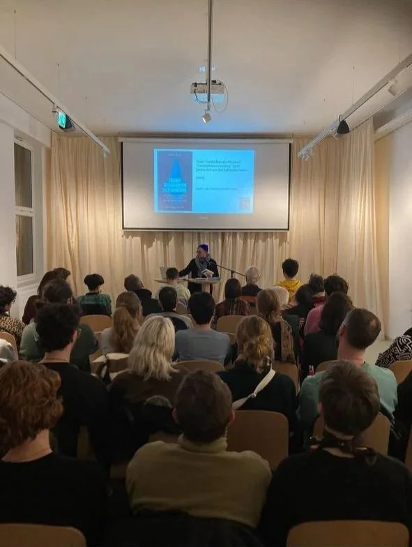 A man giving a presentation at the front of a room full of seated audience members. There is a large screen behind him displaying a slide with text and images.