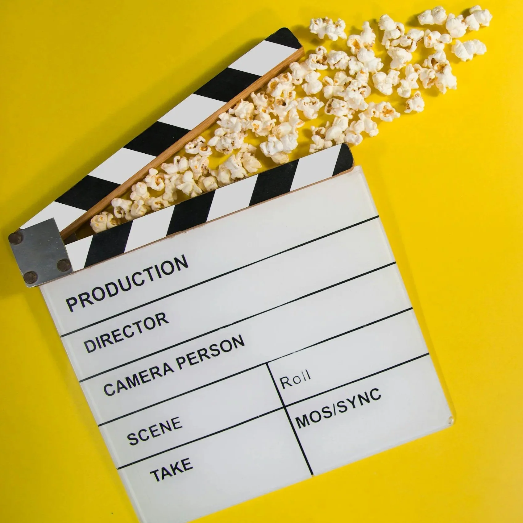 A film clapperboard with black and white stripes, partially covering a production sheet on a yellow background, with popcorn spilled onto the sheet and surrounding area.