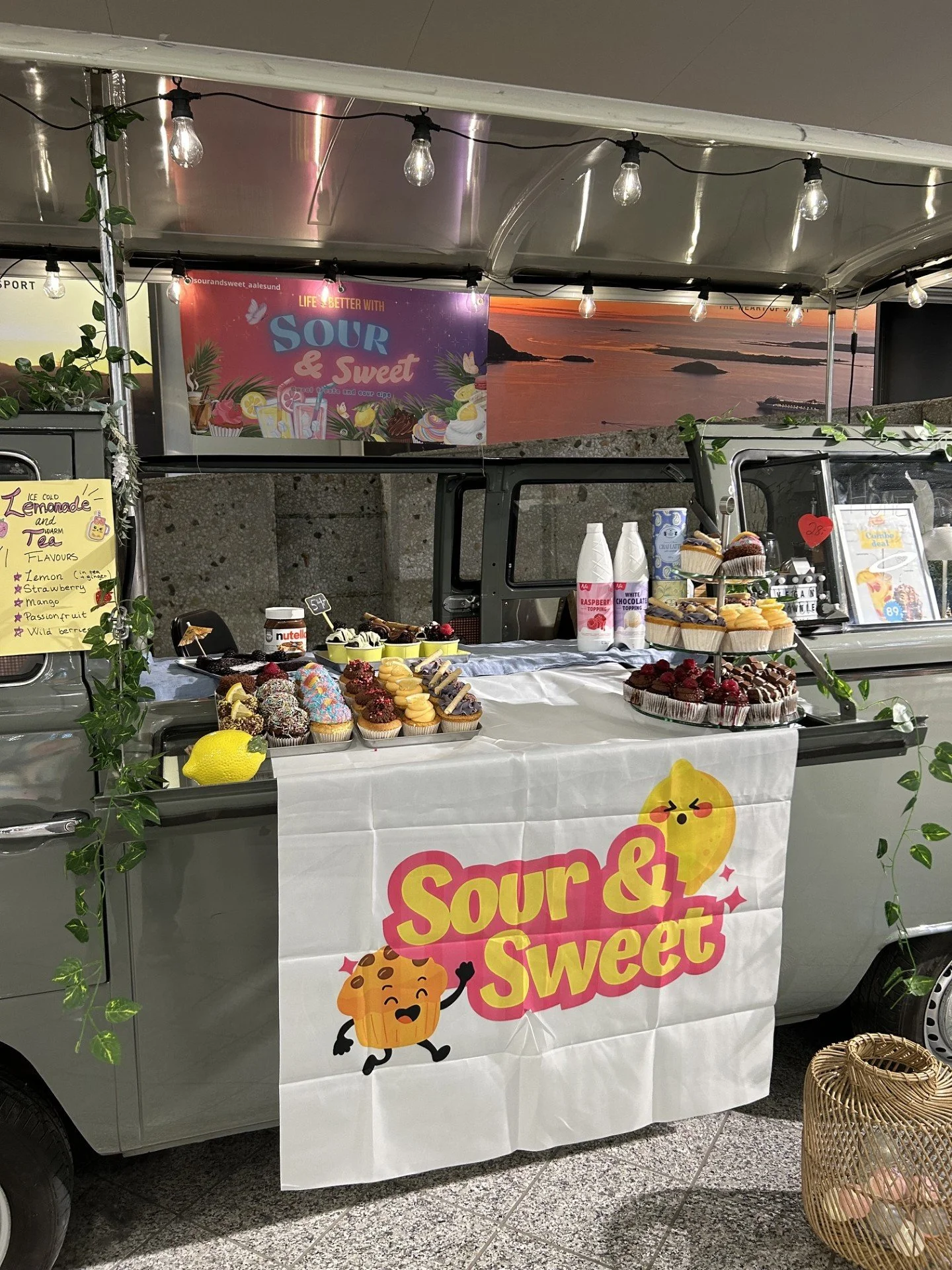 En matvogn med bakke full av muffins, kaker og desserter, dekorert med grønne blad. Det henger en plakat og en banner med teksten "Sour & Sweet" og en sitronemoji. Bak seg har et kunstutstilling med bilder av en solnedgang ved sjøen.