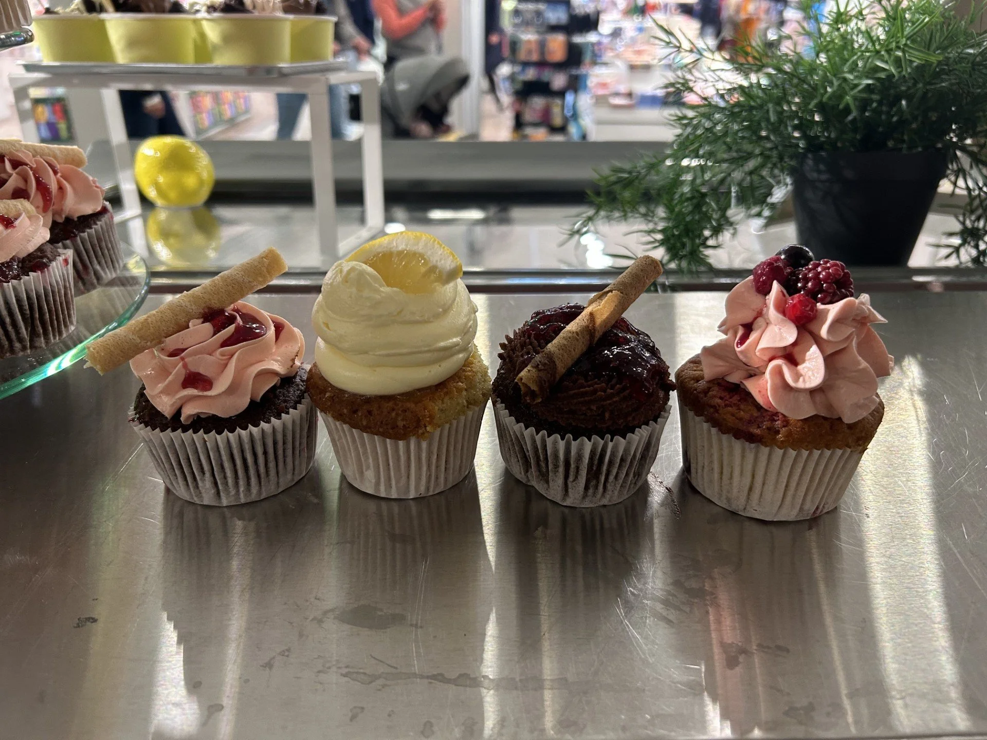 Bilde av fem cupcakes med forskjellige frosting og dekorasjoner på et metallbord i en butikk. Bakgrunnen er litt utusikkert, med mennesker og butikkhyller synlige.