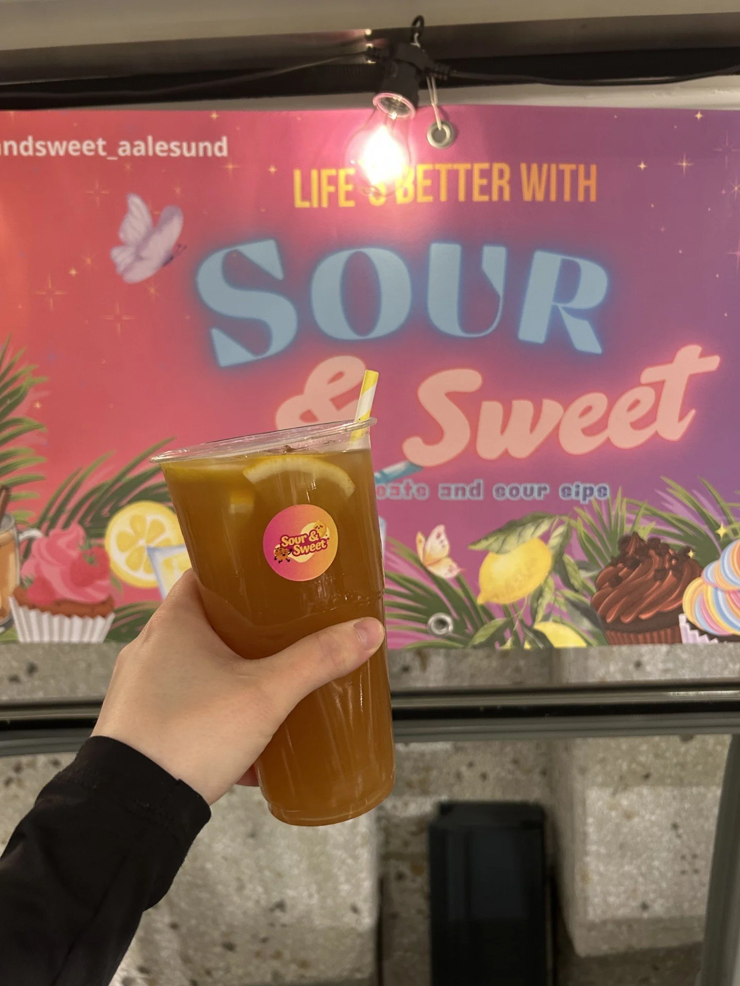 Bilde av en hånd som holder en plastkopp med en lysegul drikk med sitronbiter i foran en fargerik reklamevegg. Veggen har dekorasjoner av kaker, sitroner, blomster og en sommerfugl, med teksten 'Life's Better with Sour & Sweet'.