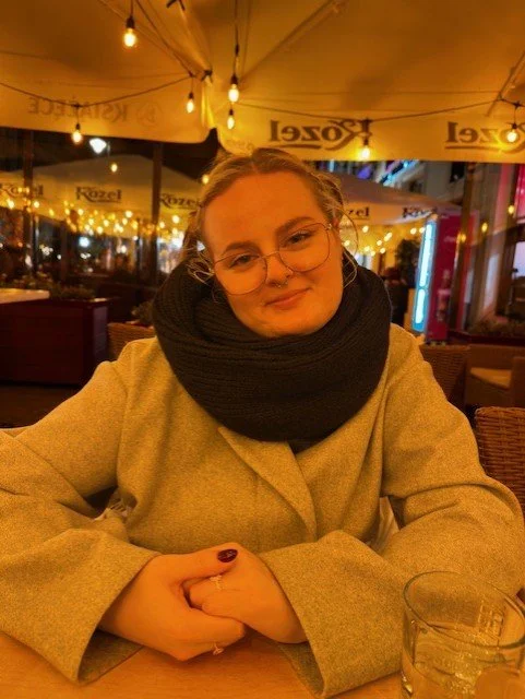 Victoria Brandal smilende, sittende ved et bord på en utendørs café om kvelden med varme lys og paraplyer med reklame. Sour and Sweet Ålesund