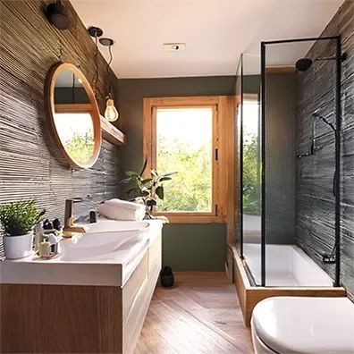 Modern bathroom with wooden framed window, green walls, dark textured accent walls, white vanity, round mirror, plant, shower with glass enclosure, and toilet.