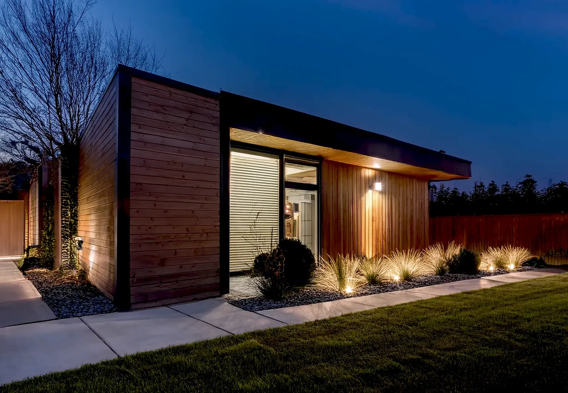A modern house with wooden exterior walls lit by outdoor lighting at dusk, featuring a flat roof, large windows, and landscaped yard with grass, shrubs, and ornamental grasses.