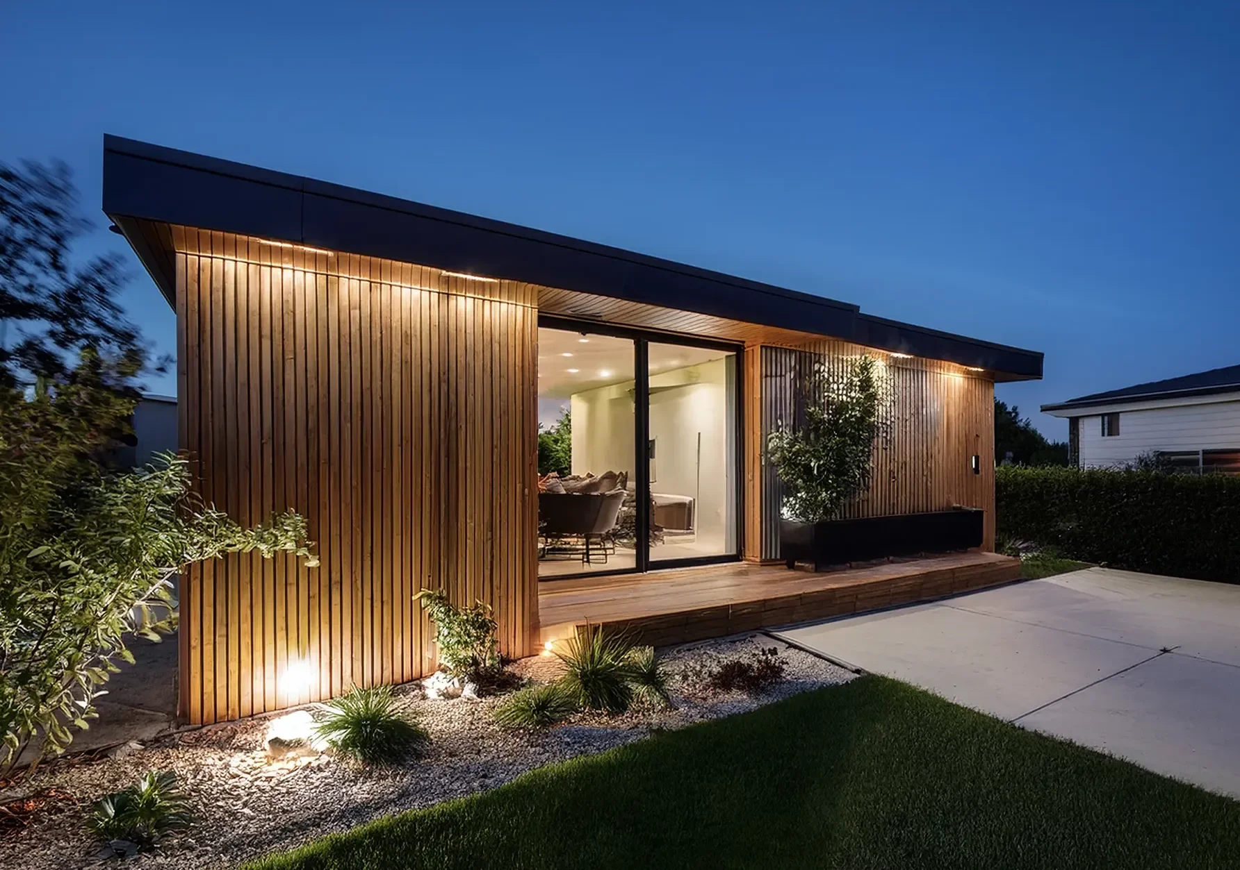 Small affordable modern ADU with wooden exterior siding, large glass sliding doors, and outdoor lighting illuminating a garden.