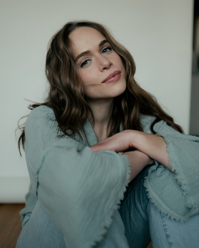 A young woman with wavy brown hair and blue eyes sitting against a plain wall, smiling softly, wearing a light gray sweater and jeans.