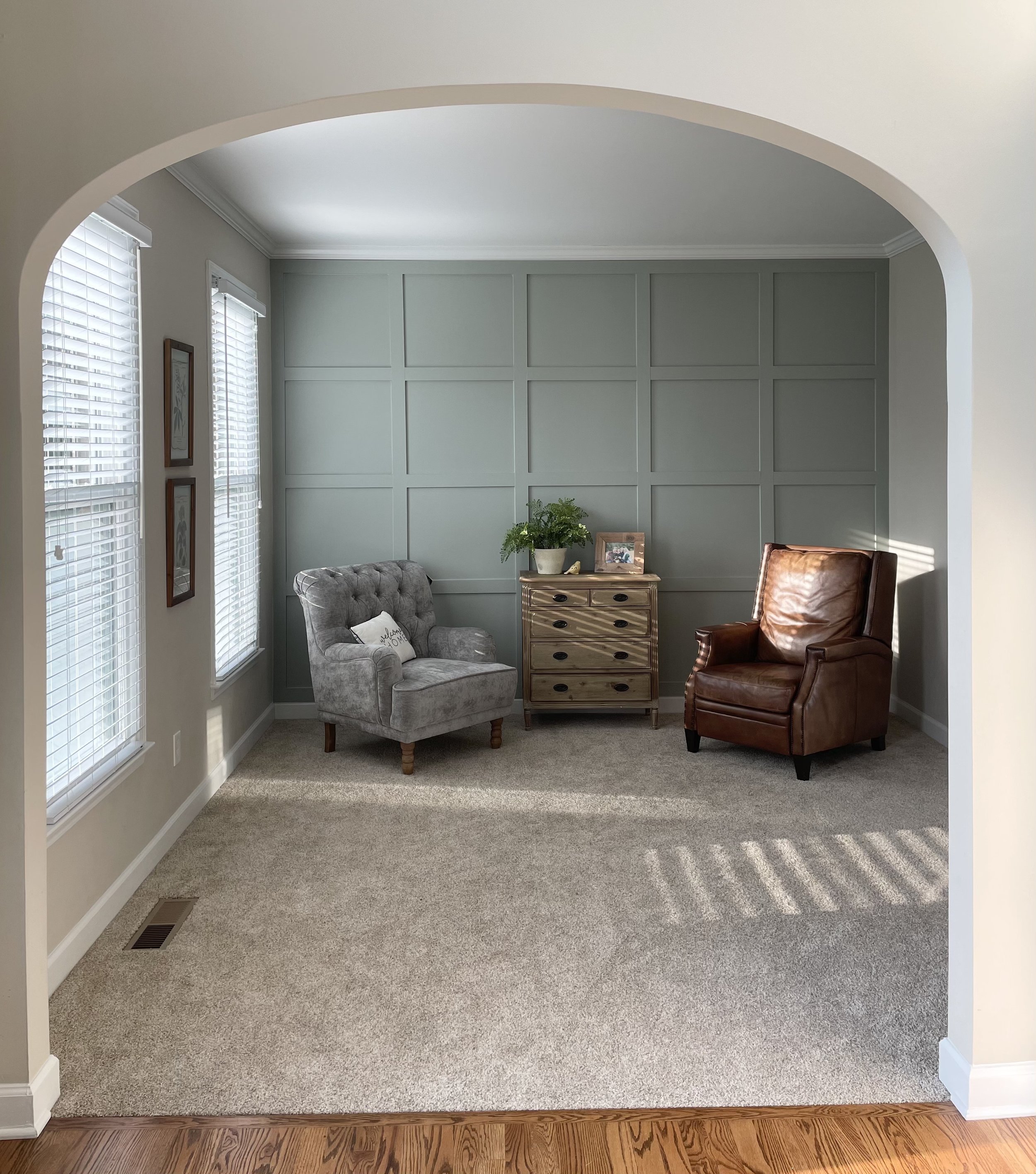 A cozy sitting area with two armchairs, one gray with a decorative pillow and one brown leather, placed on a beige carpet. A wooden side table with a potted plant, a framed photo, and small decorative items is between the chairs. Two windows with white blinds let in natural light, and a wall with green paneling is in the background.