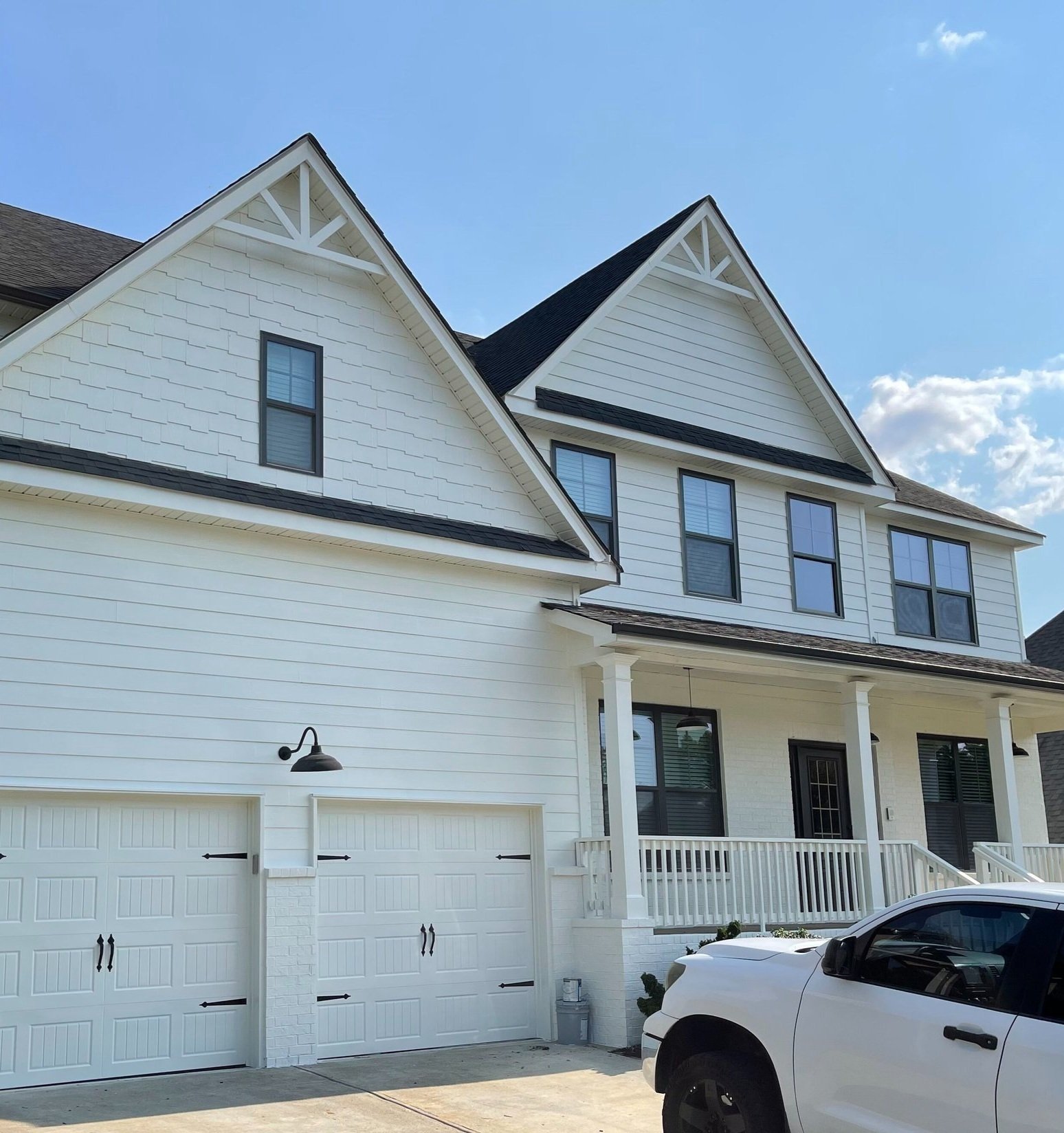 White multi-story house with black trim, attached garage, porch, and a white truck parked in the driveway, under a blue sky with some clouds.