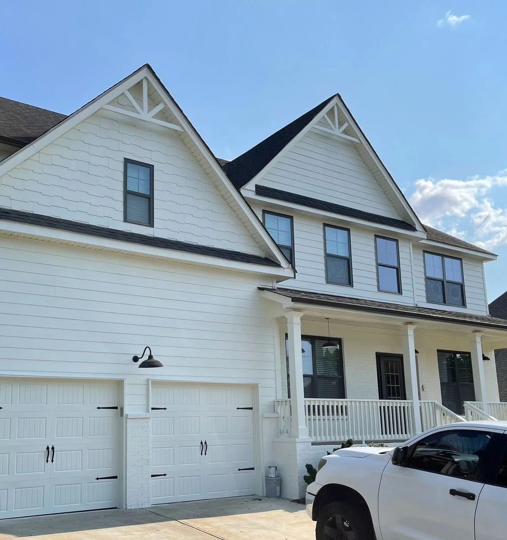 A white, multi-story house with a garage, black window frames, a front porch, and a white pickup truck parked outside under a blue sky with some clouds.
