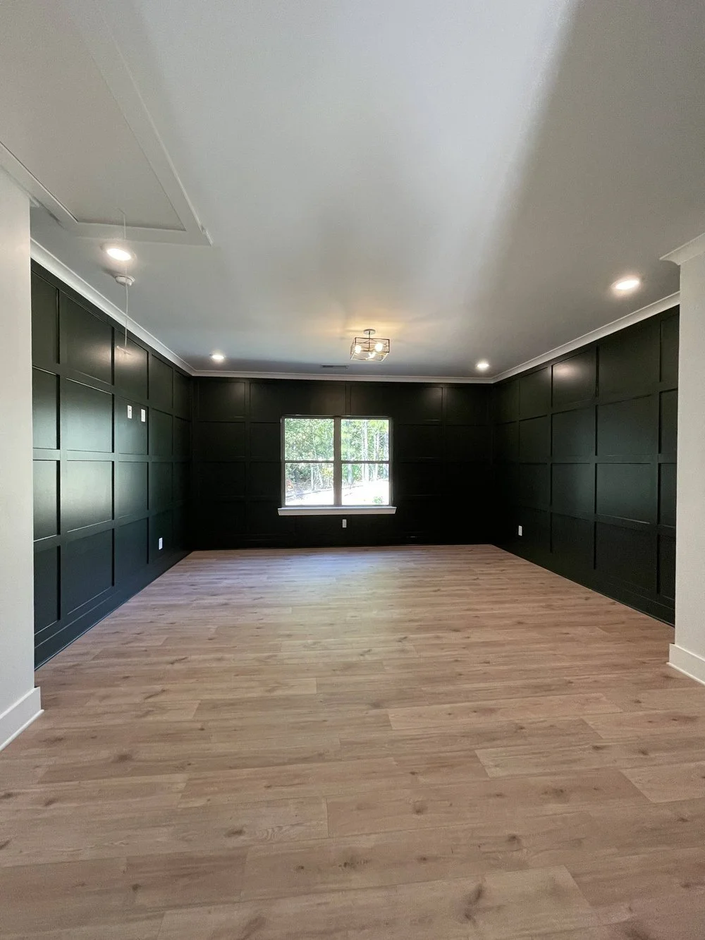 Empty living room with dark green paneled walls, light wood flooring, and a window facing outside with trees visible.
