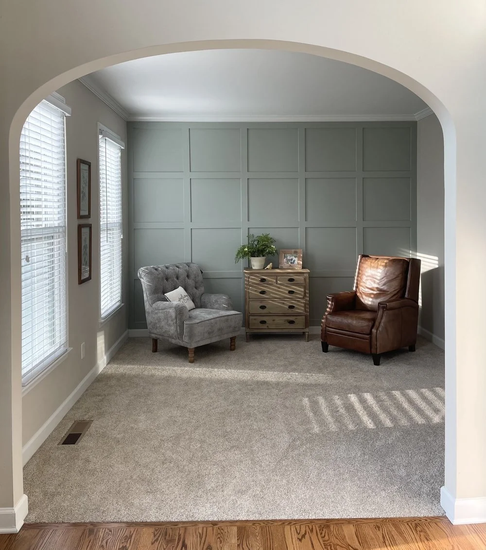 A cozy sitting area with two armchairs, one gray and the other brown, and a small wooden chest of drawers with a plant and picture frame on top, against a paneled wall and next to windows with blinds.