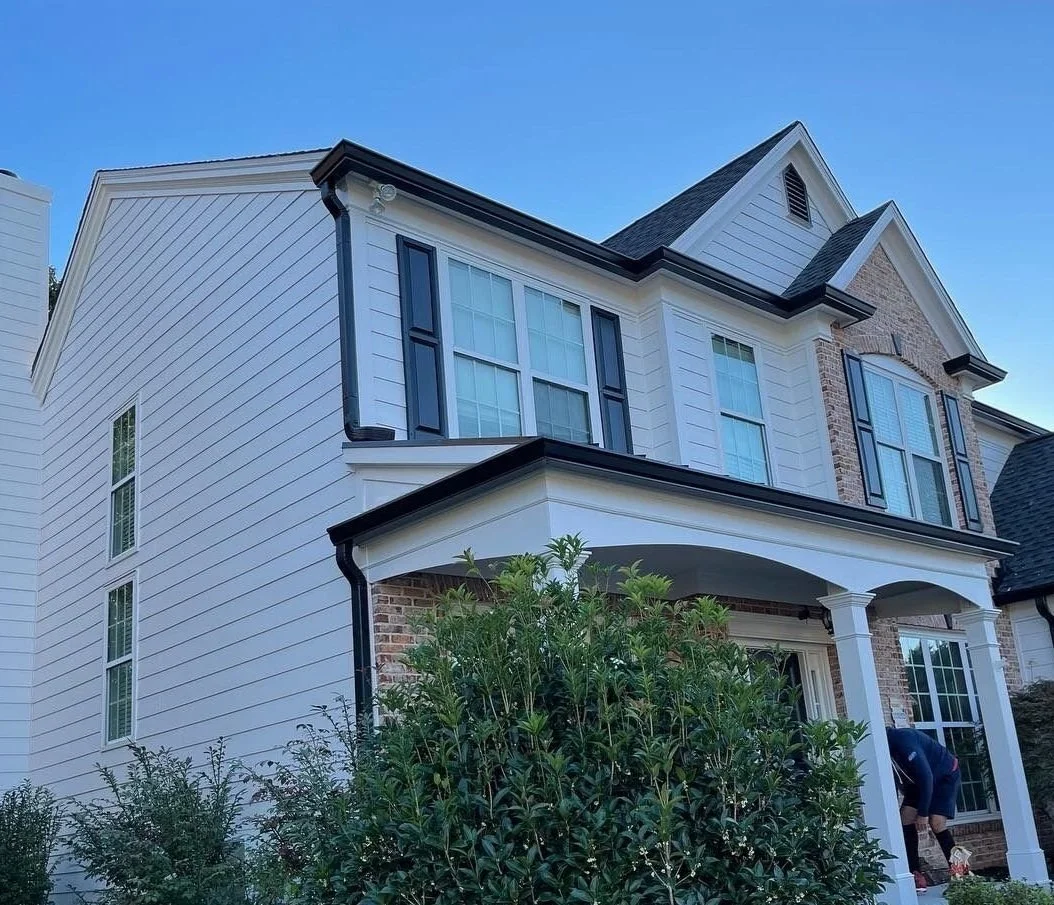 Exterior view of a two-story suburban house with white siding, brick accents, and large windows; a person is working at the front porch.