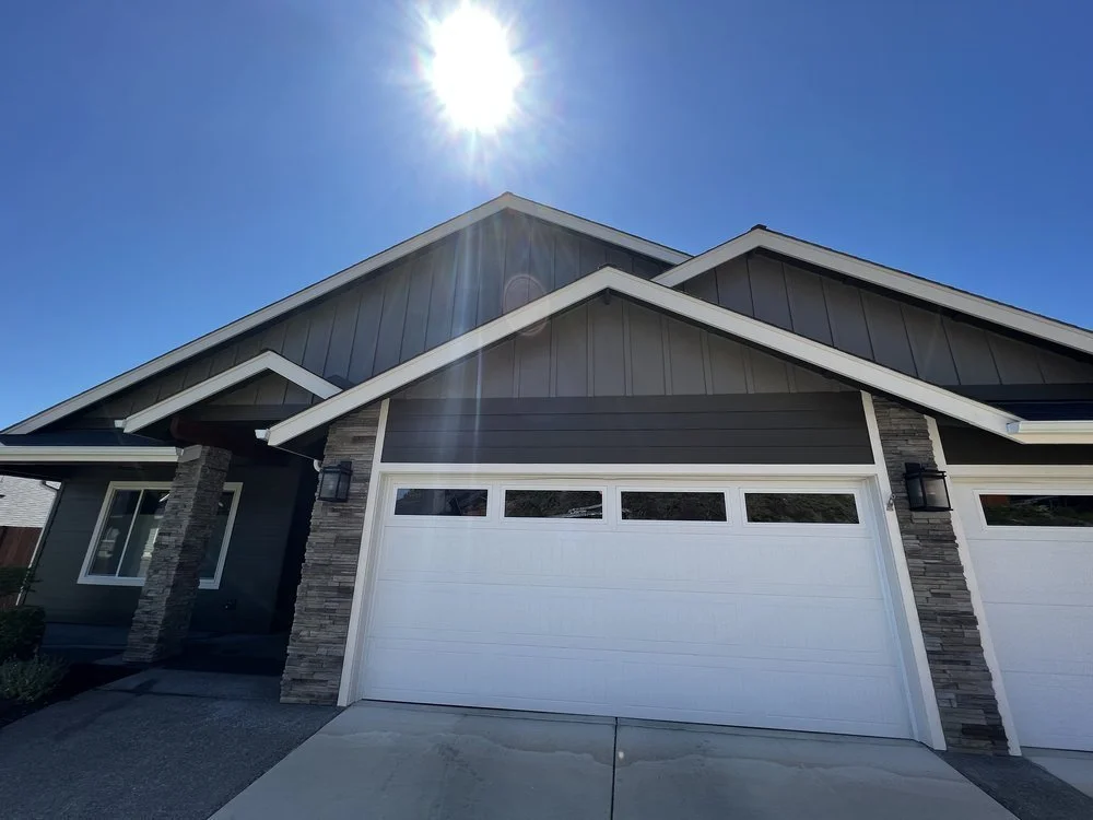 Front view of a modern house with a white garage door, stone pillars, and grey siding, under a bright blue sky with the sun shining overhead.