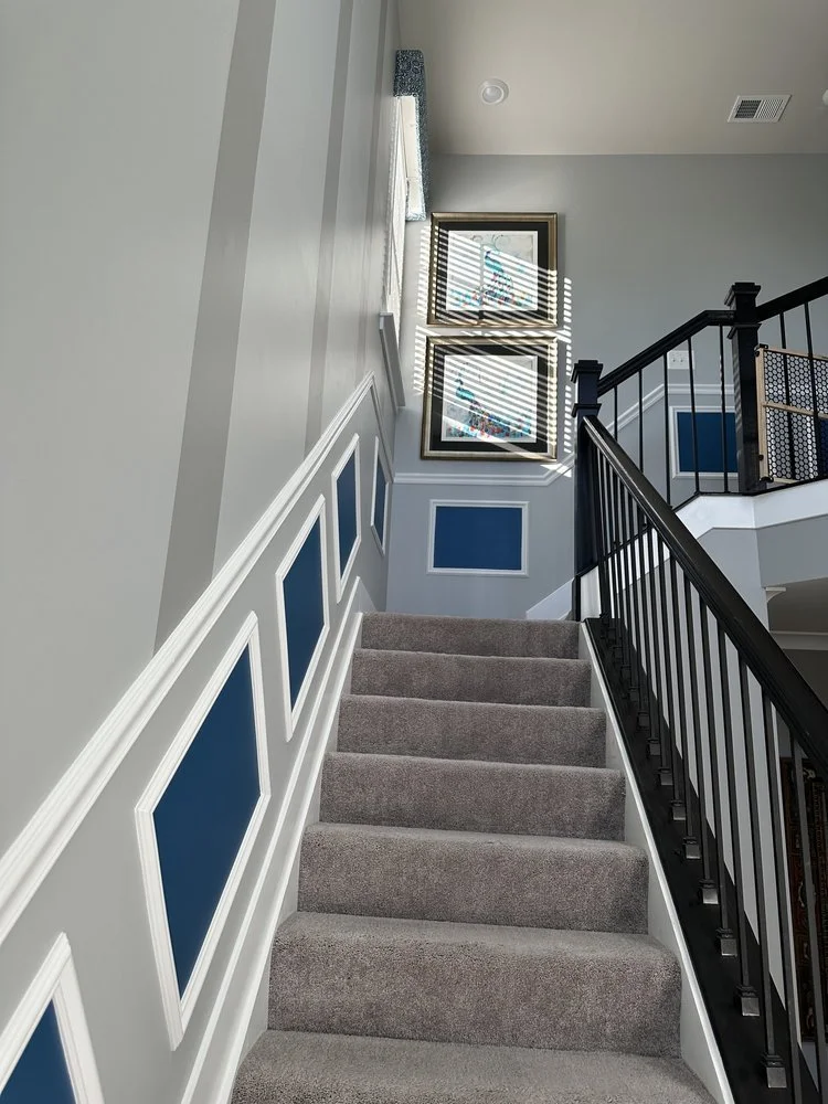 A staircase with gray carpet, white and blue wall paneling, and framed artwork near the ceiling. The area is lit by sunlight coming through a window with blinds, casting shadows on the wall.
