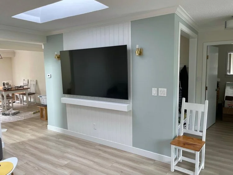 Living room with a large flat-screen TV mounted on a light green wall, a white shelf below the TV, a white wooden chair with a brown seat to the right, and a dining area with a table and chairs in the background.