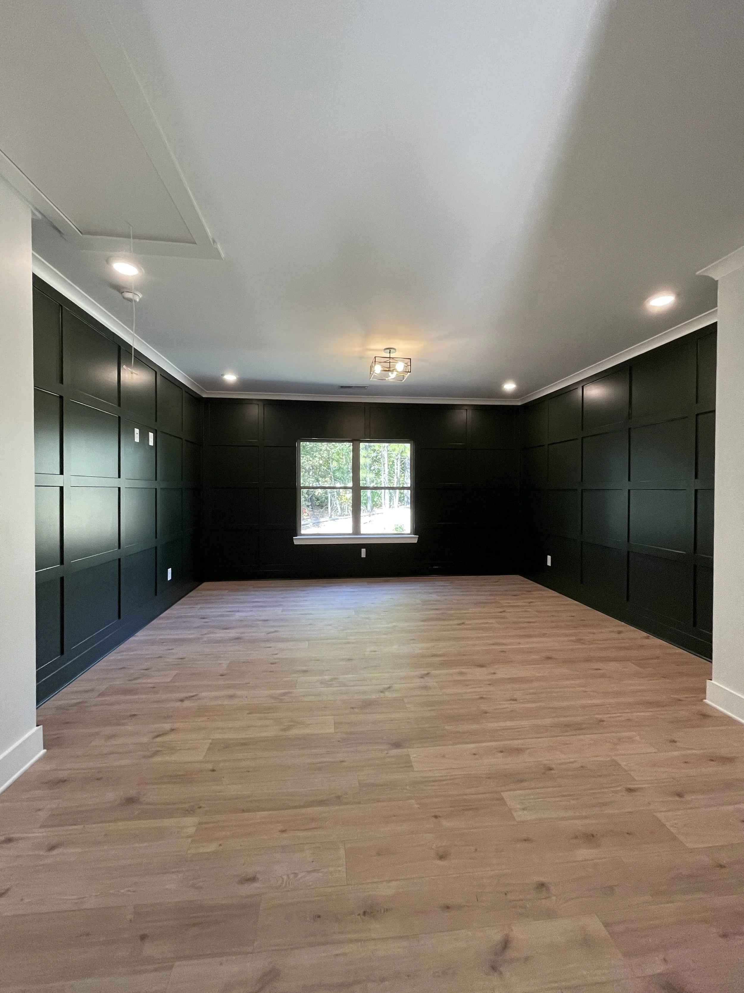 Empty room with black paneled walls, light wood flooring, a window showing trees outside, and ceiling lights.