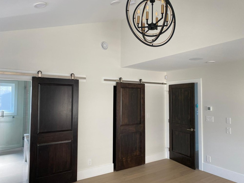 Interior of a room with dark wooden sliding barn doors, white walls, a black chandelier, and a small window in a bathroom visible through an open door.
