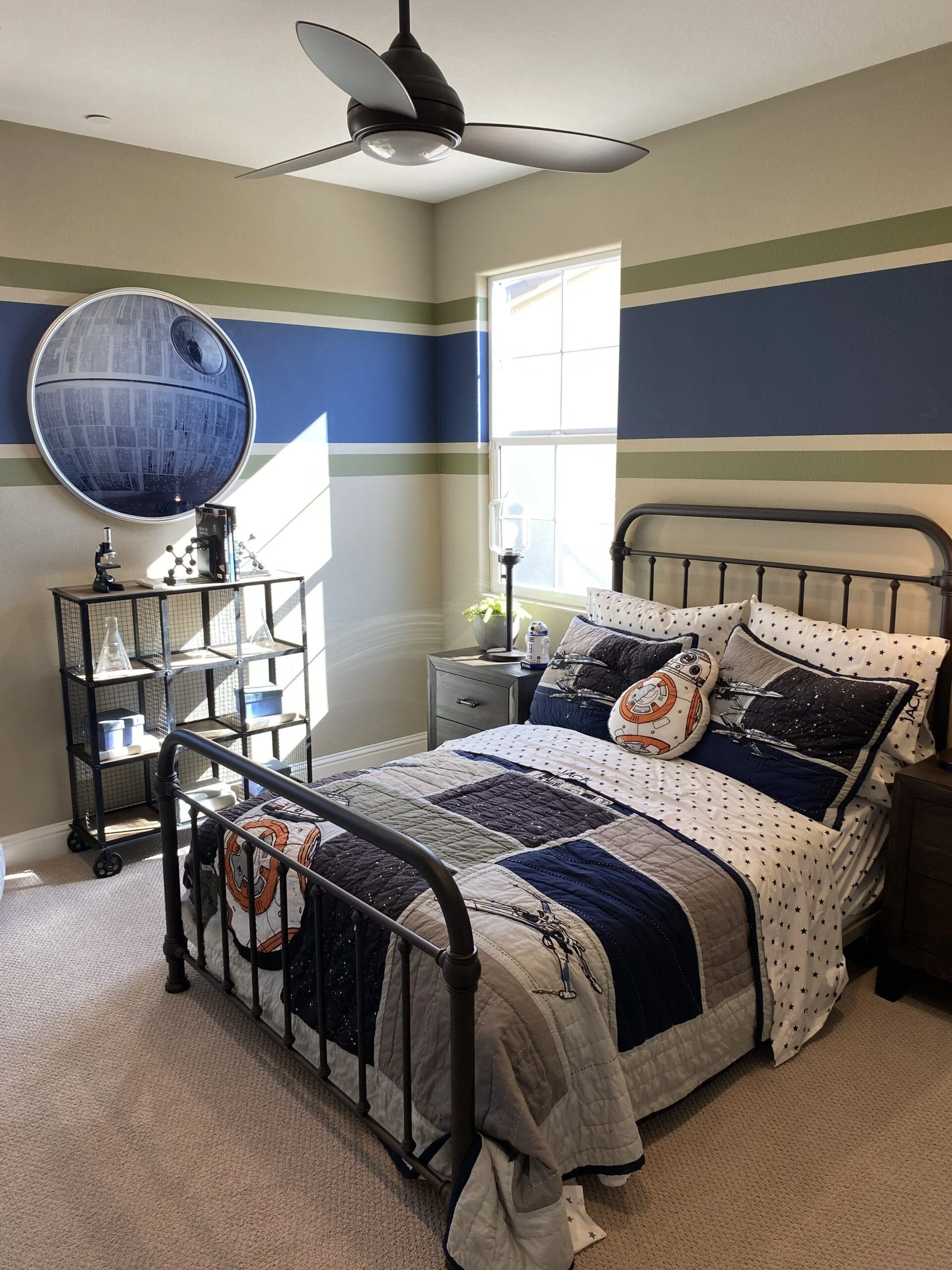 A bedroom with beige walls and blue and green striped accents. There is a metal bed with Star Wars-themed bedding featuring BB-8. Next to the bed is a dark wood nightstand with a water lamp and a small plant. A black shelving unit holds various items