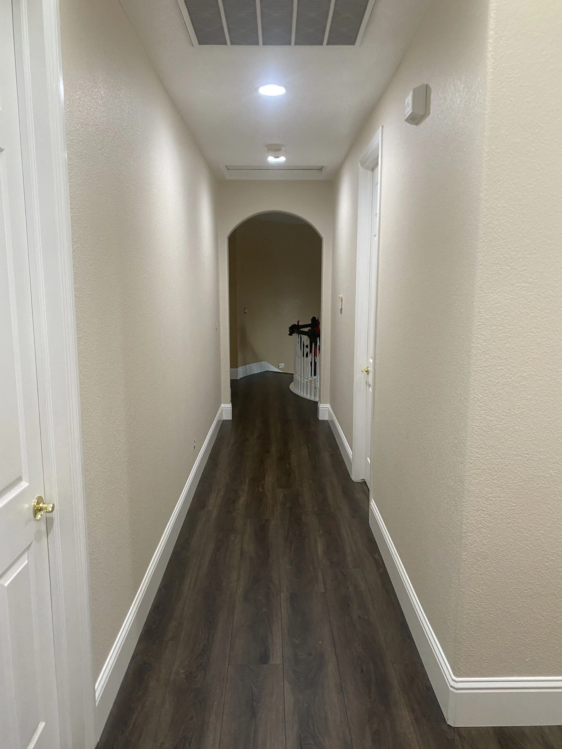 Empty hallway in a house with beige walls, white baseboards, a dark wood floor, and a ceiling vent, with a staircase visible at the end.