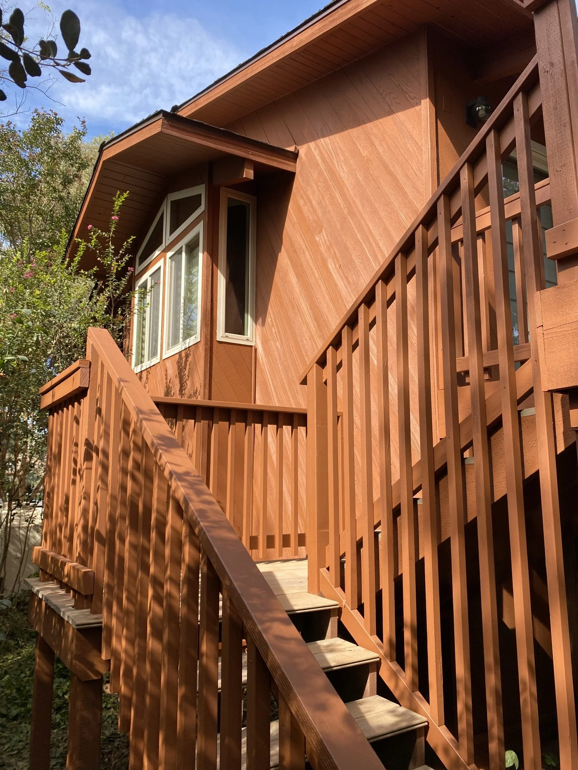 A wooden house with a staircase and railing, and a window with a geometric design, surrounded by trees and clear blue sky.