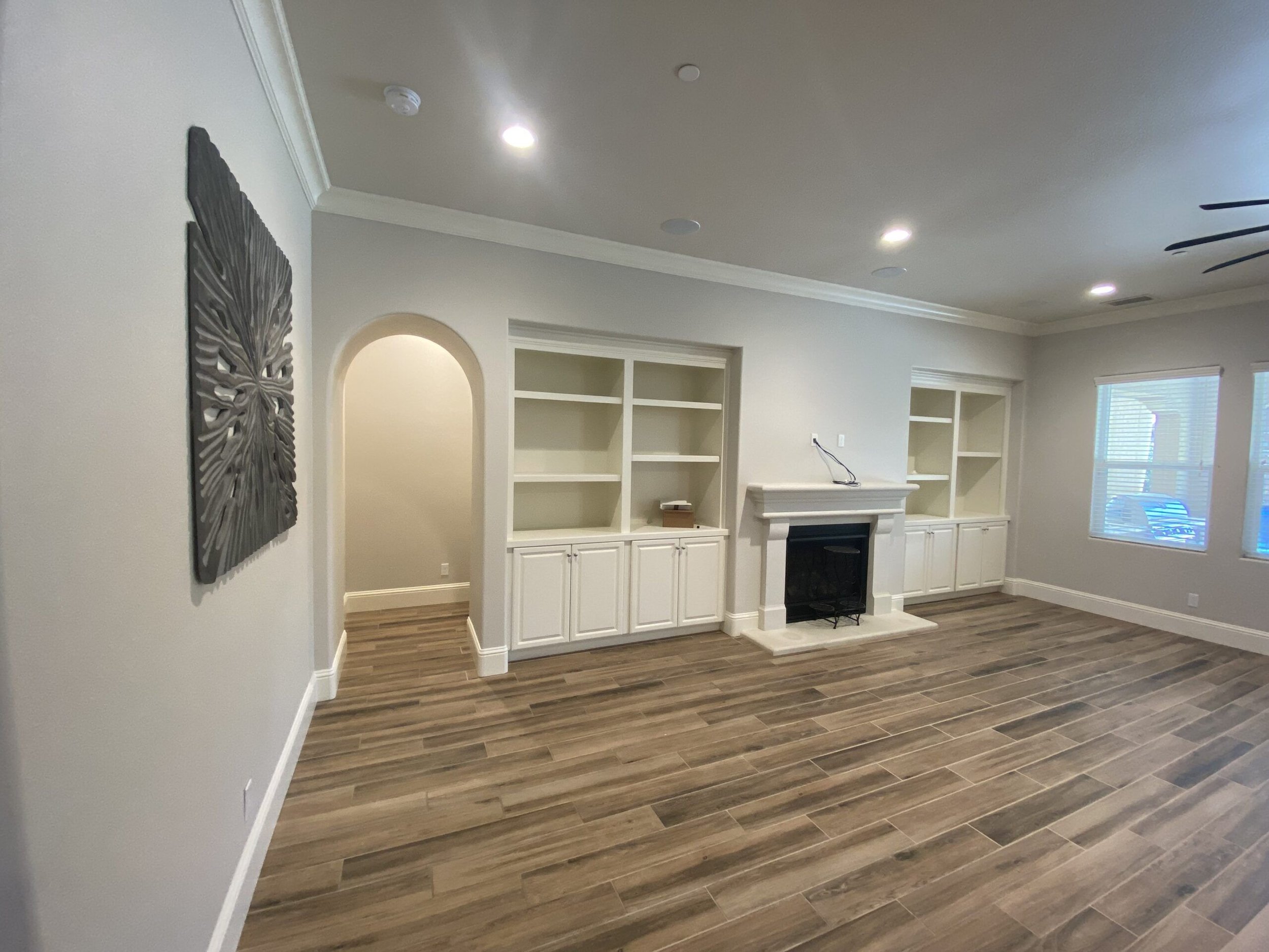 Empty living room with white built-in shelves around a fireplace, wood-look flooring, and a wall art piece on the left wall.