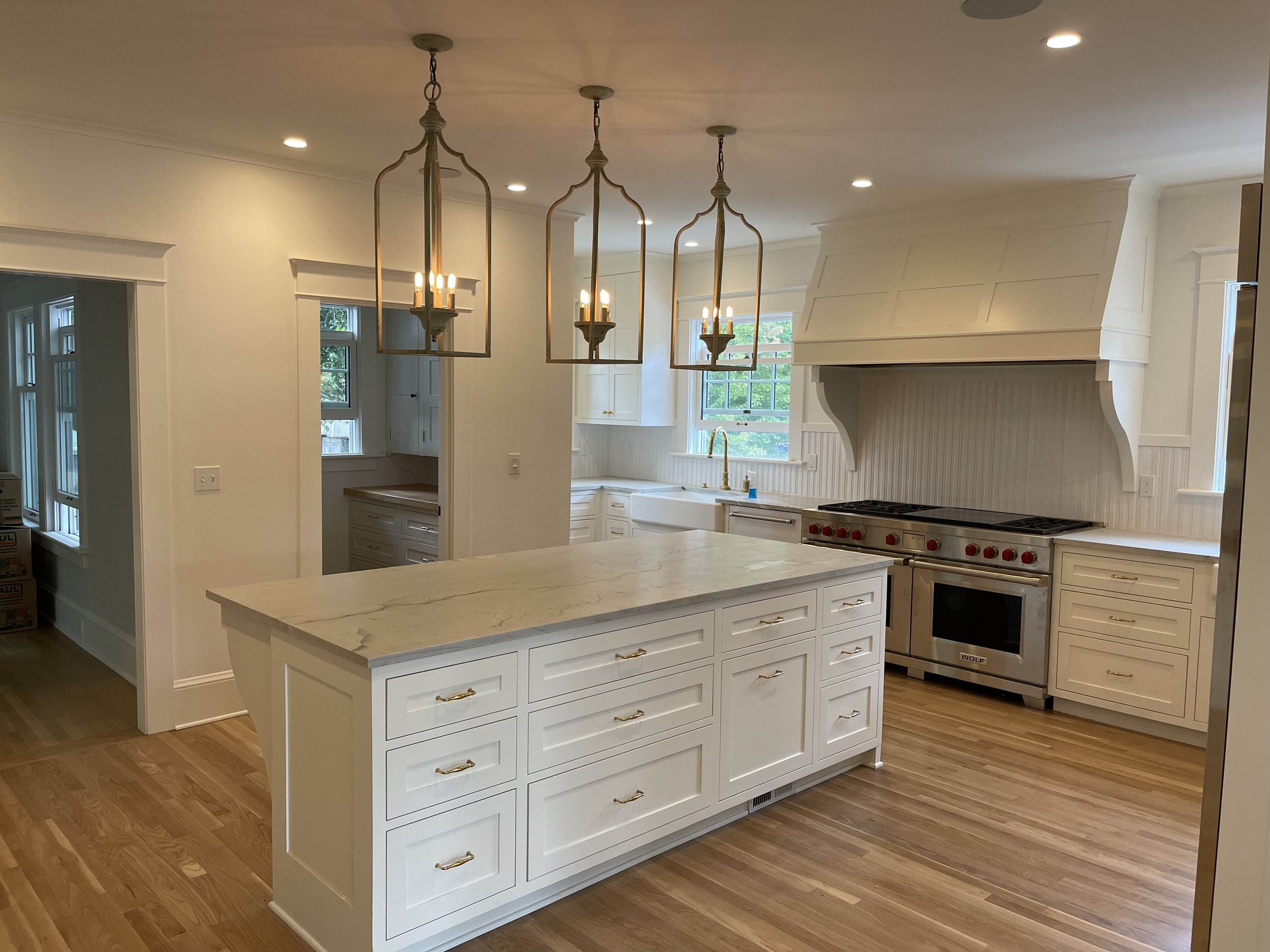Modern white kitchen with central island, brass hardware, hardwood floors, and a large stove with red knobs under a white range hood. Pendant lights hang over the island, and there are cabinets and windows with natural light.