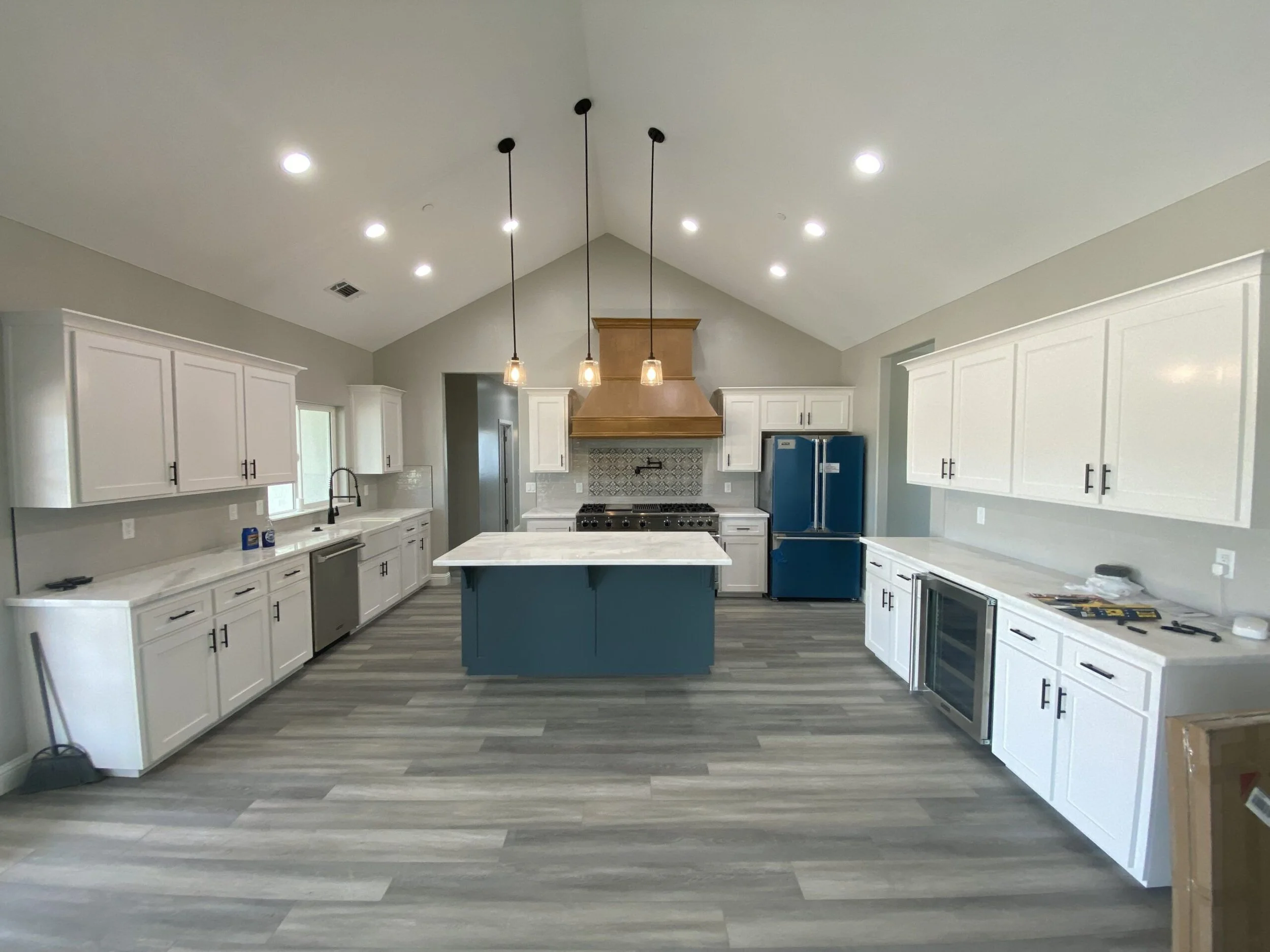 Modern kitchen with white cabinets, a blue kitchen island, stainless steel appliances, and a gray and white tiled backsplash beneath a vaulted ceiling with recessed lighting and pendant lights.